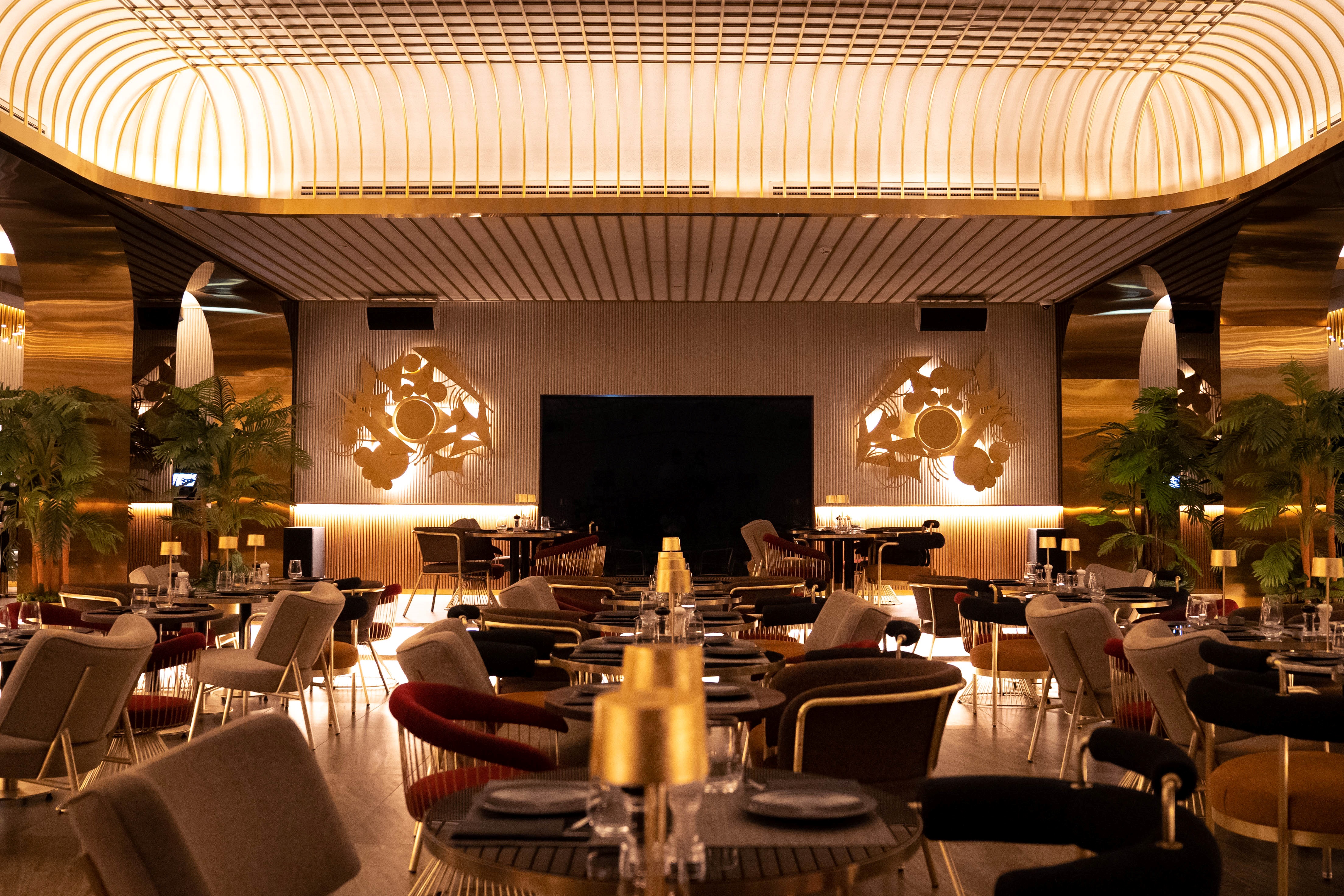 Modern restaurant interior with gold accents, stylish chairs, round tables, and warm lighting.