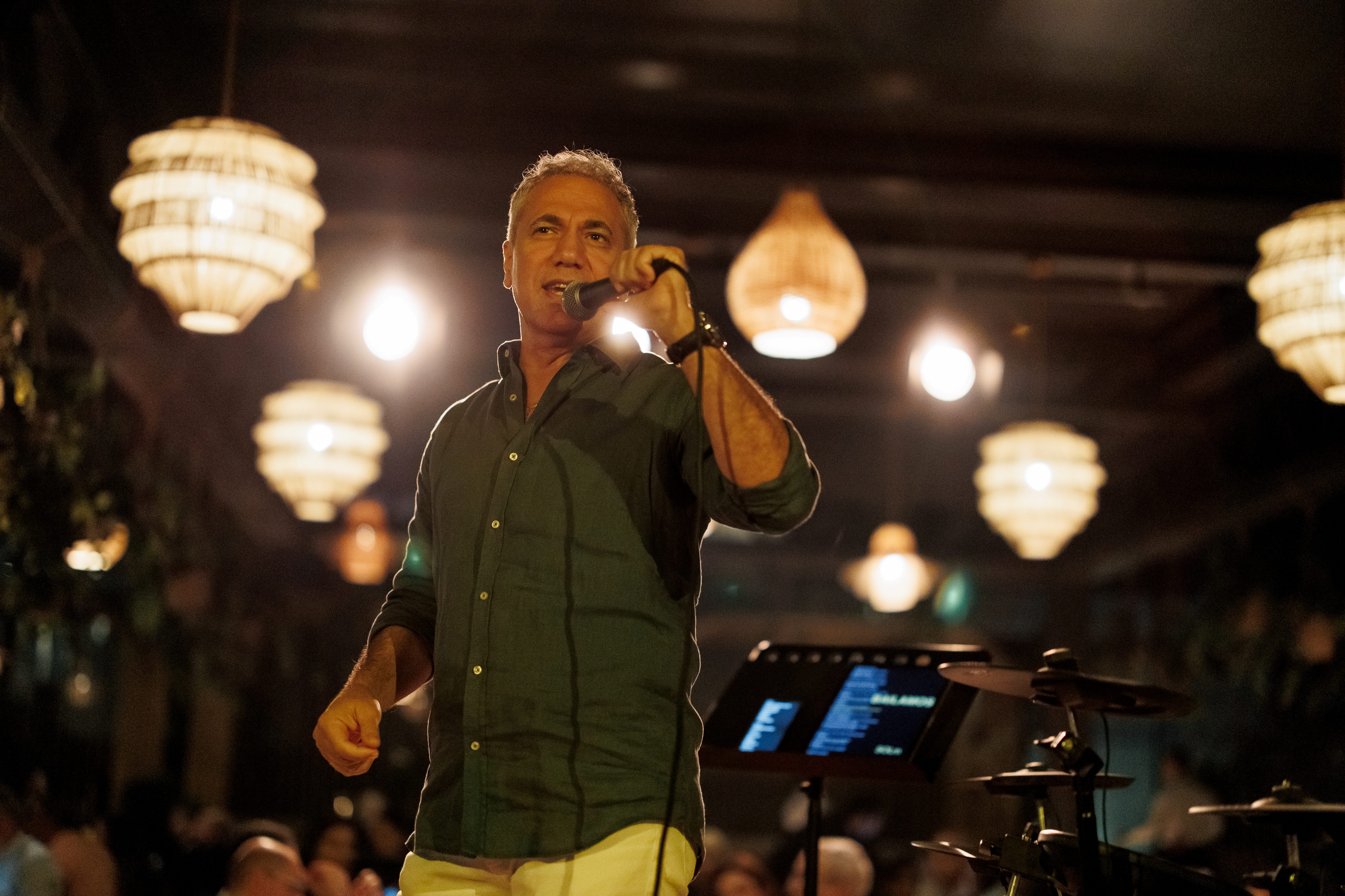 Man in green shirt singing into a microphone on stage with hanging lights in the background.