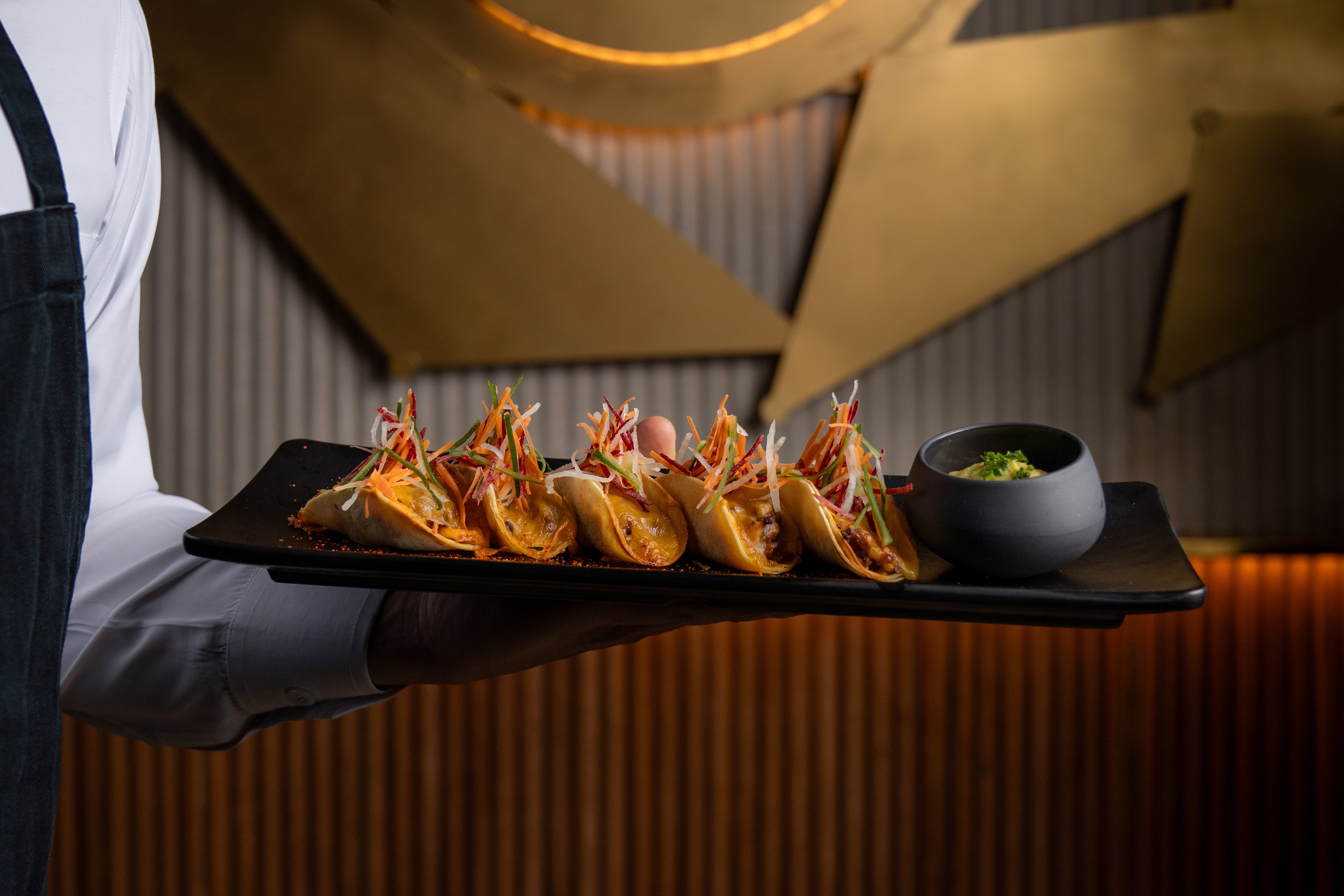 Server holding plate with five small tacos topped with shredded vegetables and a side of dipping sauce.