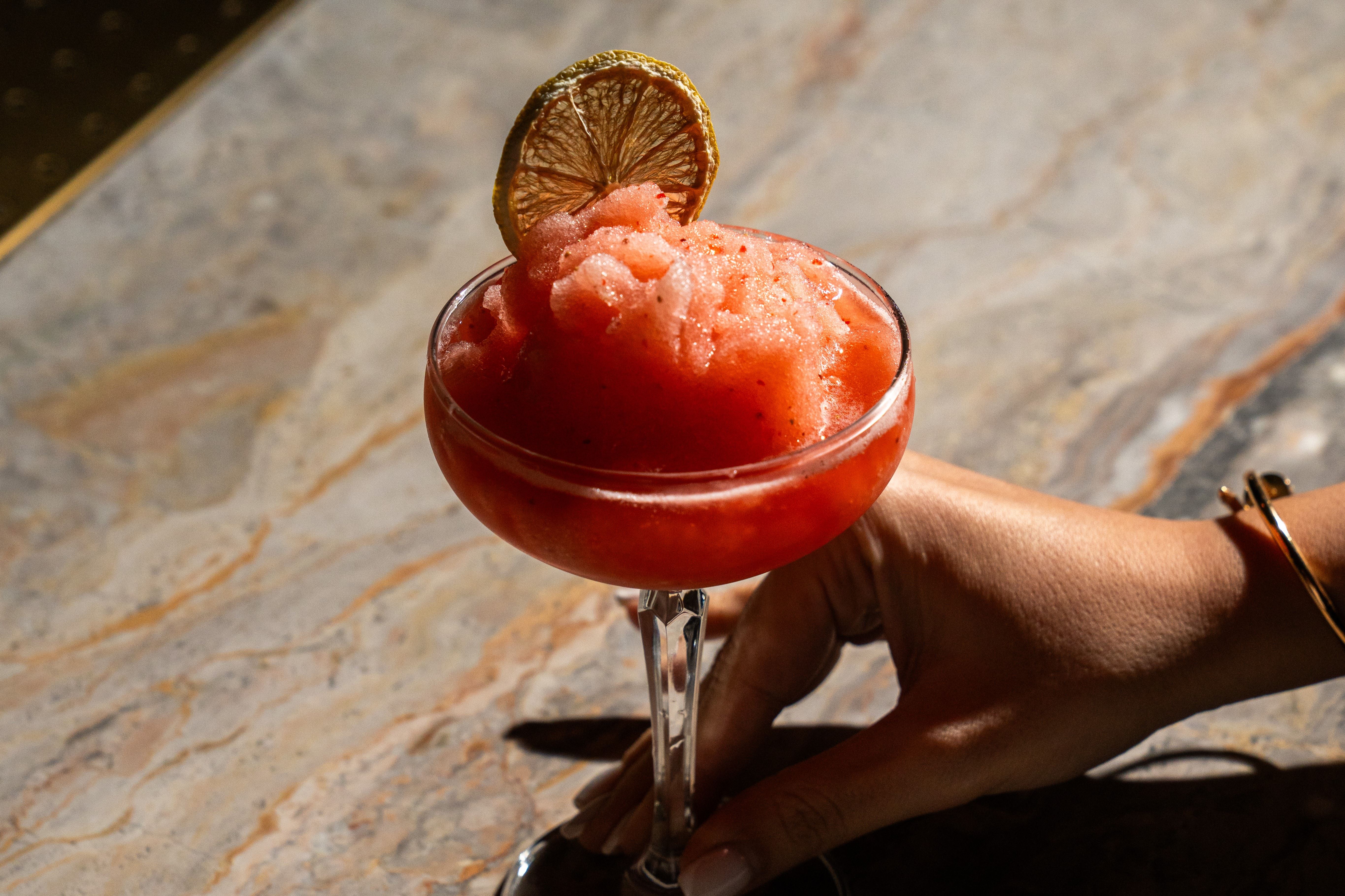 Hand holding a glass of red frozen cocktail garnished with a dried lime slice on a marble surface.