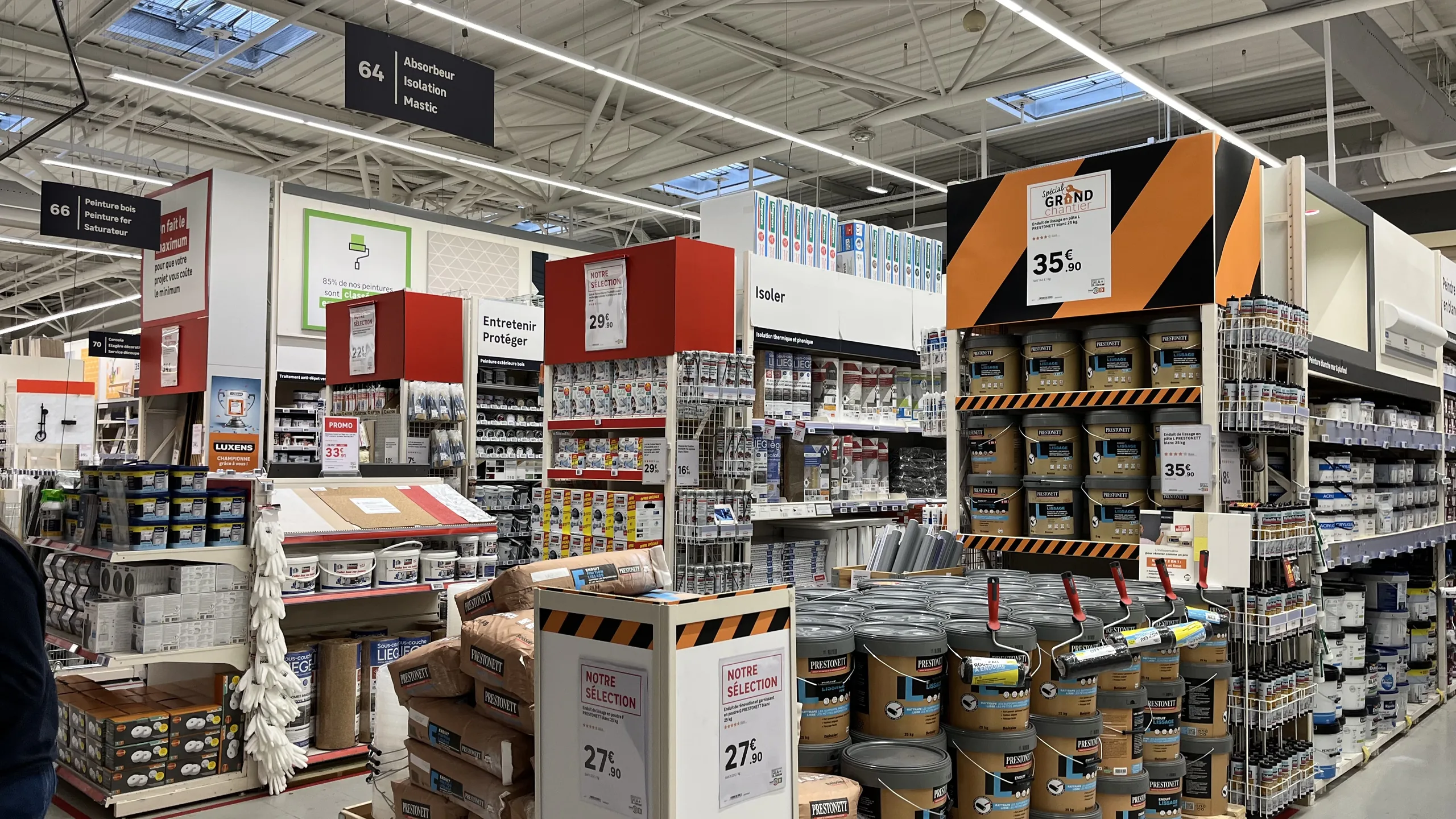 Allée principale d'un magasin de bricolage bordée de têtes de gondole avec frontons de couleur et affiches prix