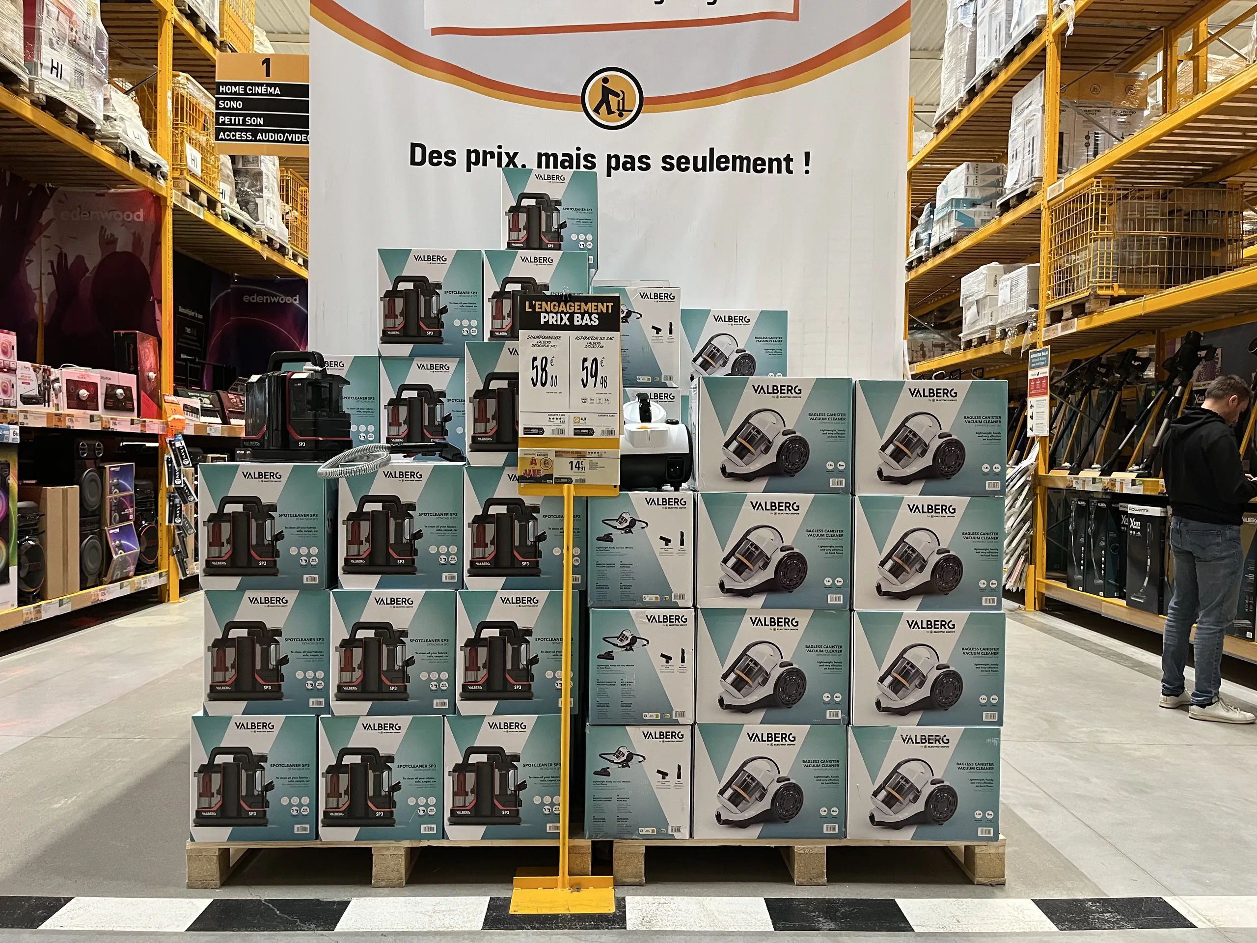 Palette de produits d'électroménager en allée de magasin, avec une grande bannière de message et une affiche prix sur pied