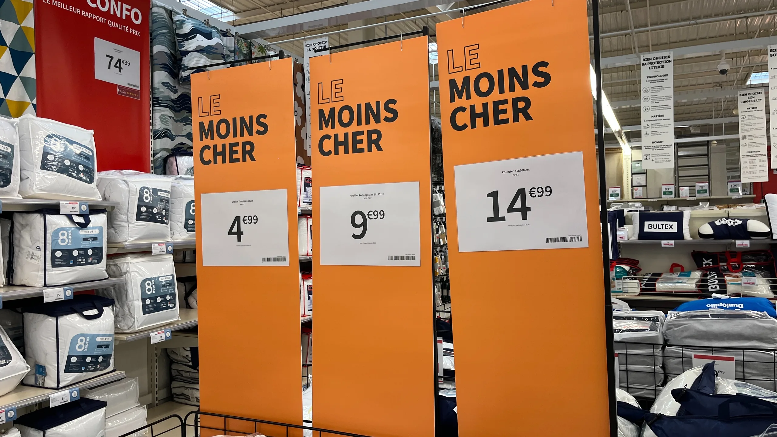 Trois kakémonos orange « Le moins cher » suspendus au-dessus d'un rayon de linge de lit, chacun avec son affiche prix