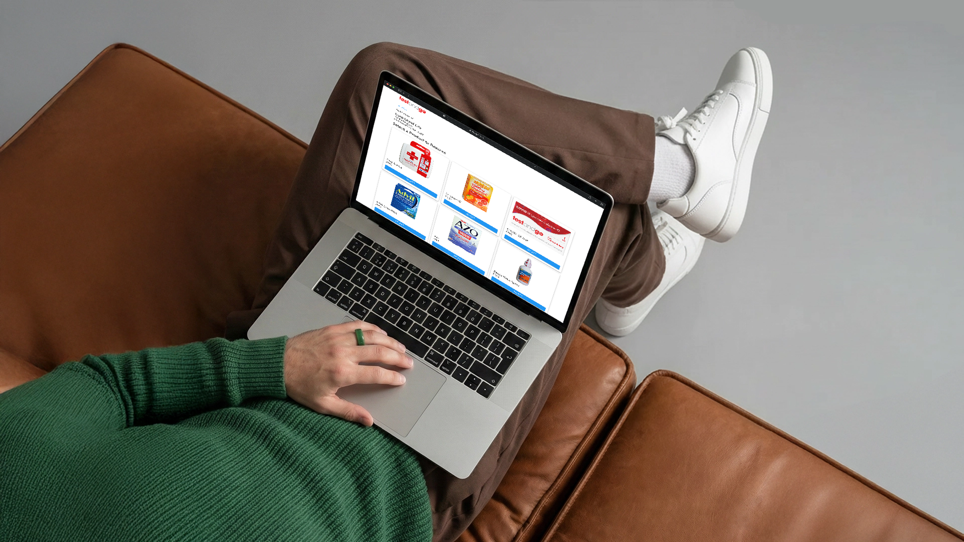 Person in green sweater using a laptop showing a webpage with product thumbnails on a brown couch.
