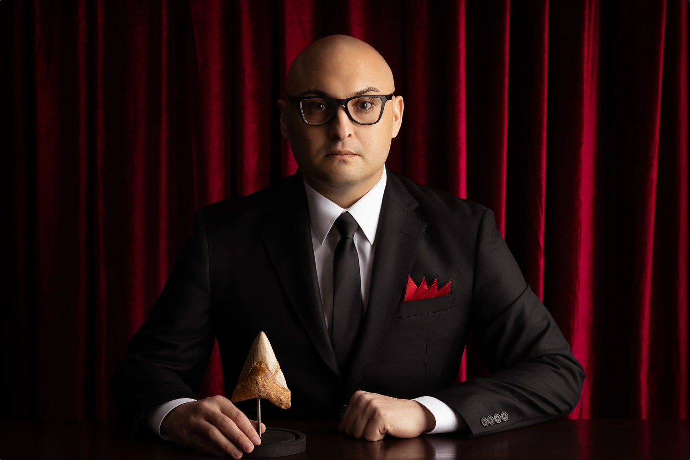 Richard Adame seated in a black suit before red curtains with a Megalodon tooth display, representing leadership and professional experience.