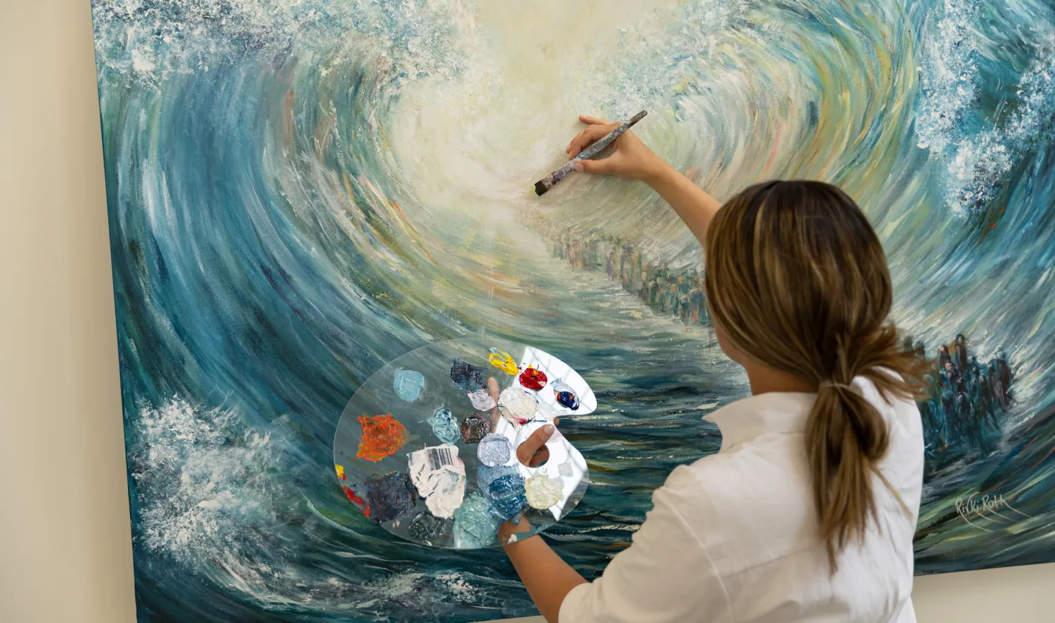 Woman with ponytail painting a large, swirling ocean wave scene on canvas while holding a palette with various paint colors.