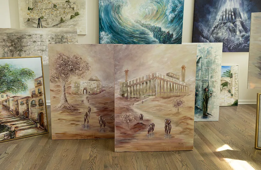 Multiple paintings displayed on a wooden floor featuring landscapes, architectural buildings, and abstract scenes, with muted and earthy tones.