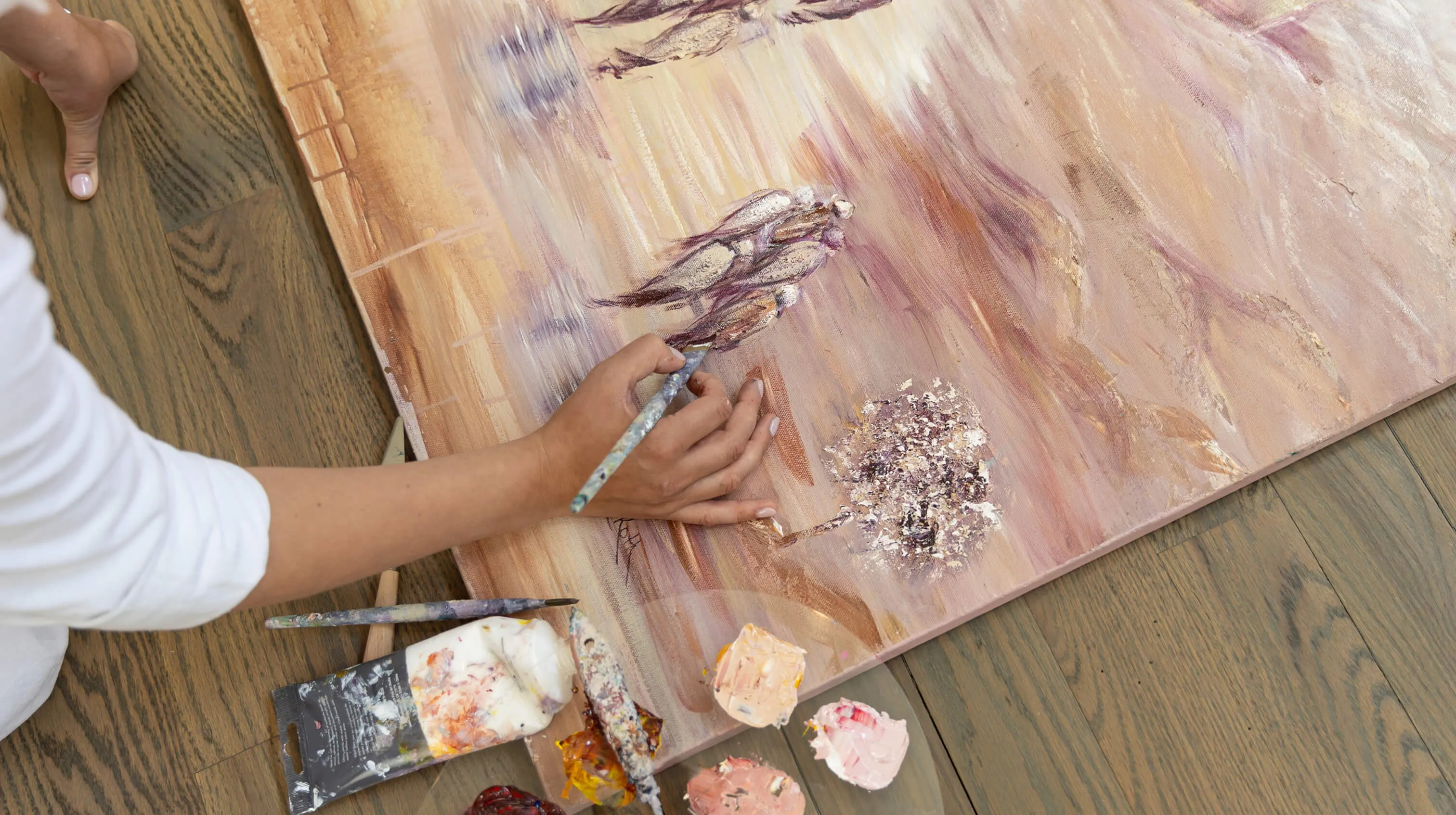 Person painting a textured artwork with figures and a tree using a brush and palette on wooden floor.