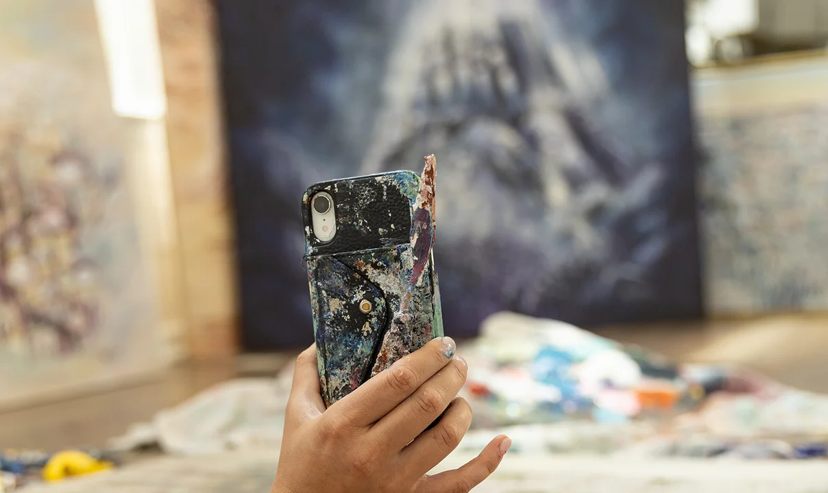 Hand holding a paint-covered smartphone case with an artist's work area blurred in the background.