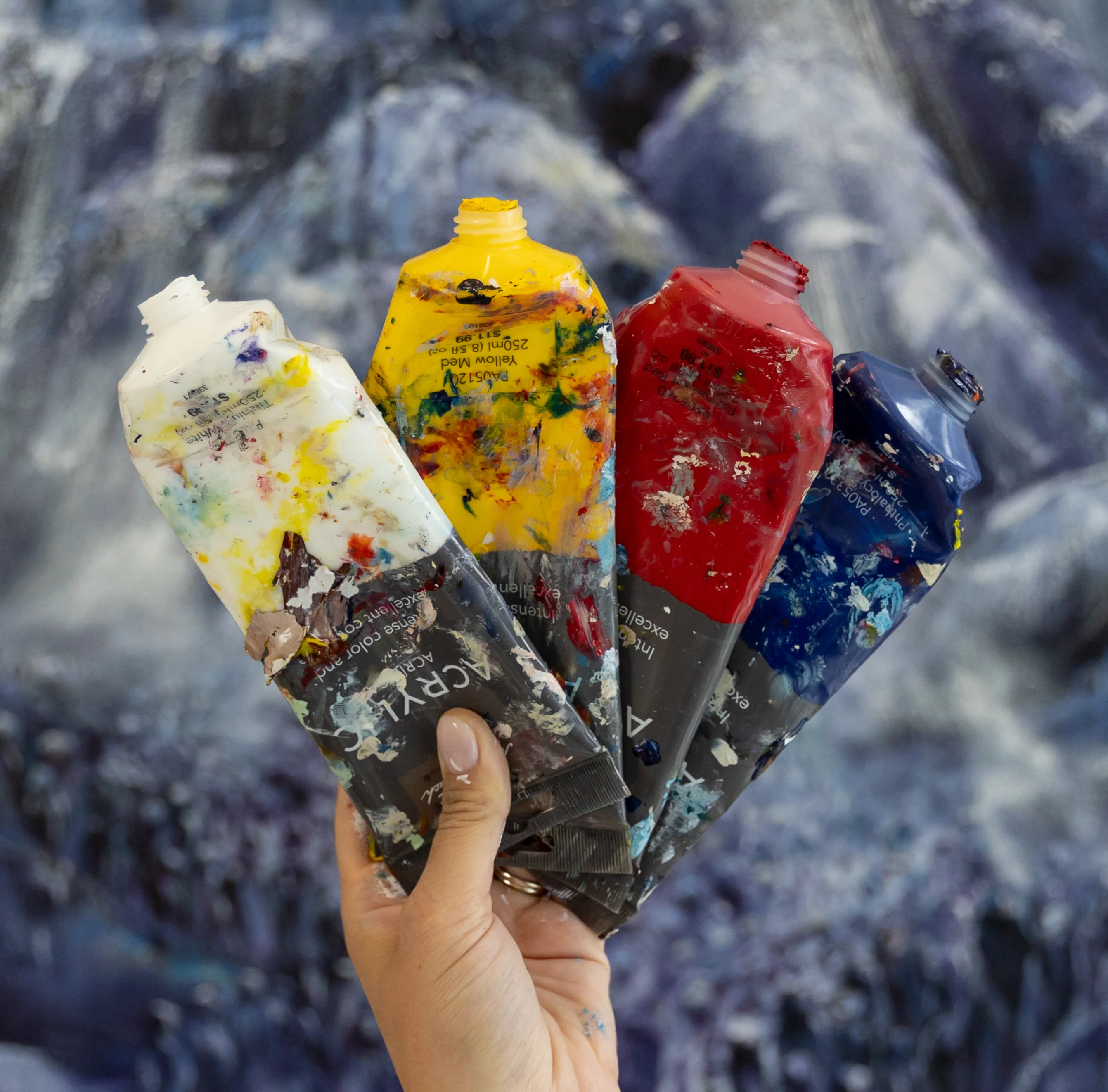 Hand holding four used acrylic paint tubes with white, yellow, red, and blue paint, against a blurred artistic background.