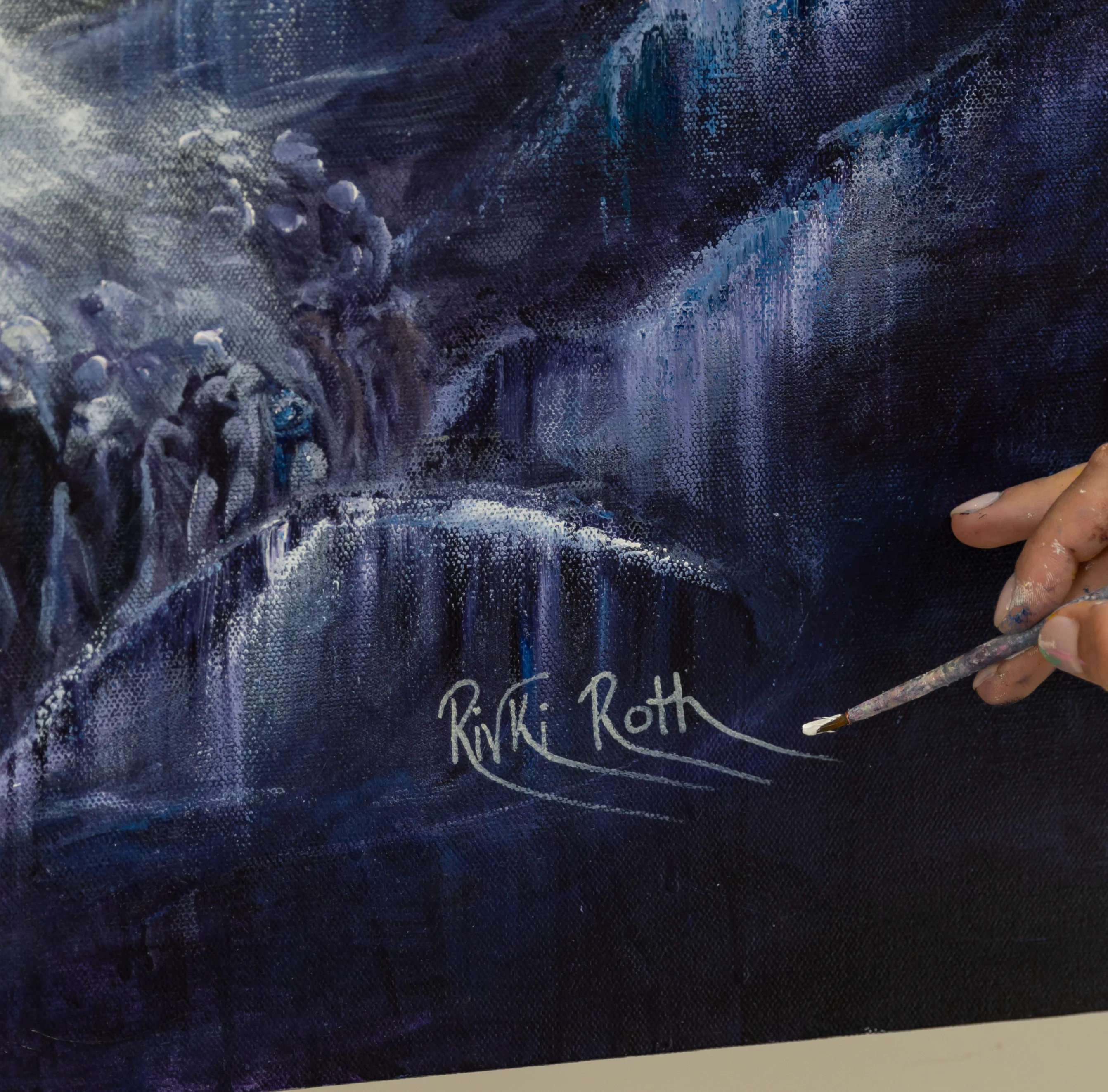 Close-up of a dark blue and purple painting featuring waterfall-like textures, with a hand holding a paintbrush near the artist's signature 'Rivki Roth'.
