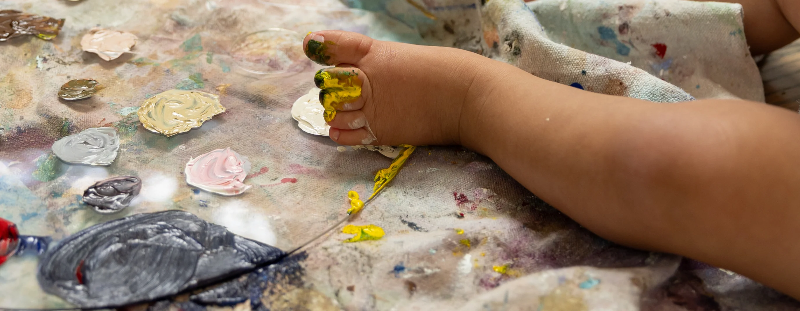 Child’s foot covered in yellow paint resting on a canvas with dabs of various paint colors.