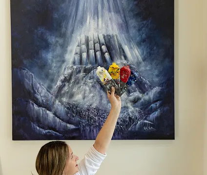 A person holds up colorful paint tubes beneath a dramatic painting of rays shining down on a dark, mountainous abstract landscape.