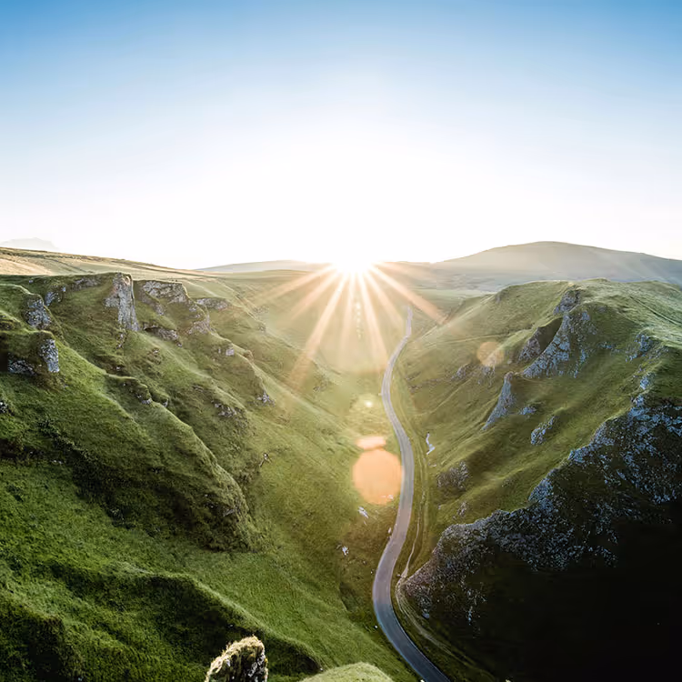 Sunrise over a winding road through green hills and rocky cliffs.