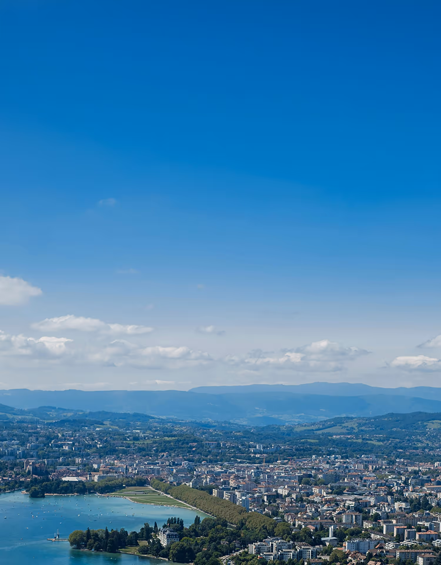Aerial view of Geneva showing Lake Geneva with boats, city buildings, tree-lined parks, and distant hills under a blue sky with scattered clouds.