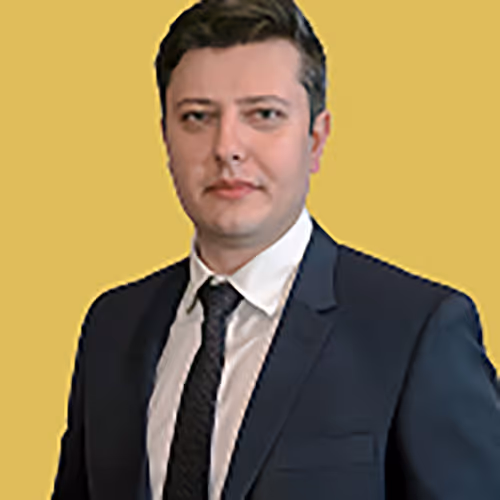 Man with short dark hair in a black suit, white shirt, and black tie against a yellow background.
