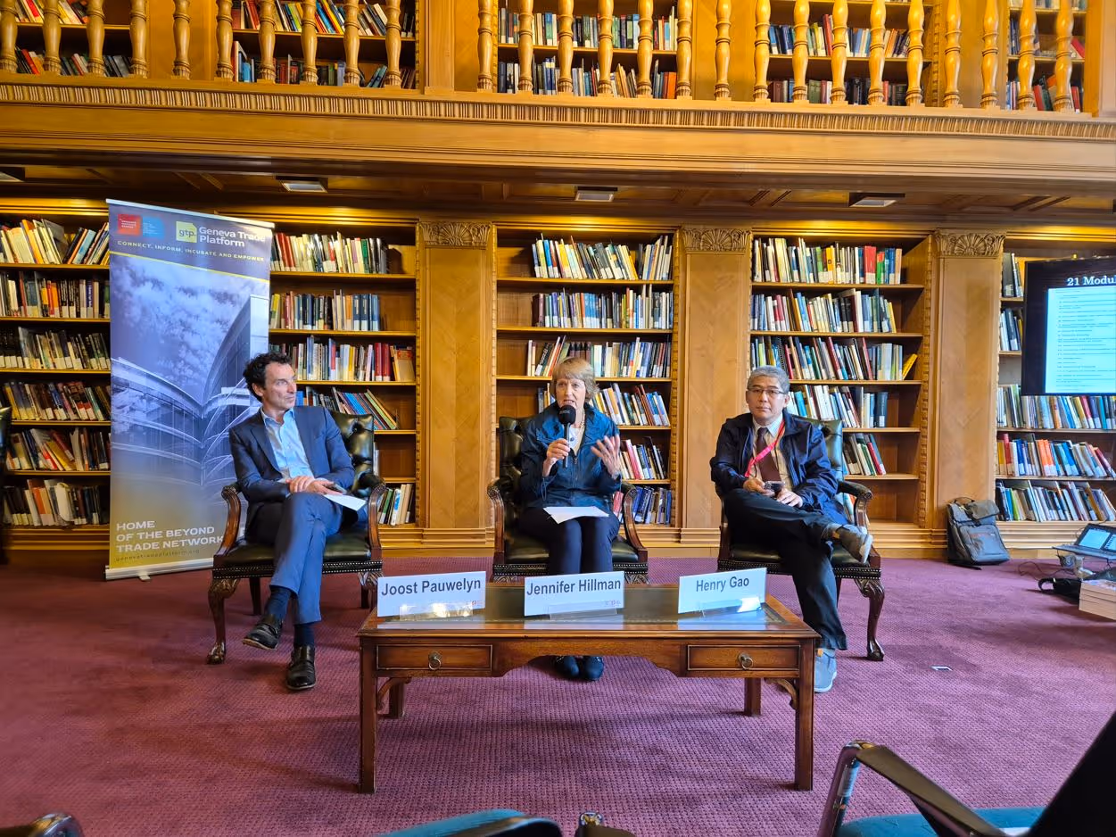 Three panelists seated in a library-style room with bookshelves, discussing with name tags in front.