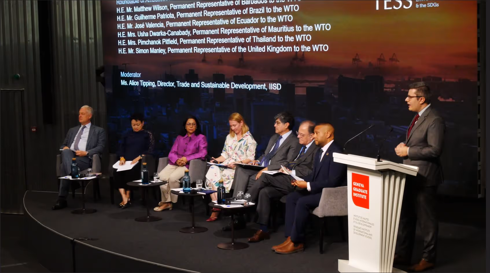Seven panelists seated on stage with one standing and speaking at Geneva Graduate Institute podium.