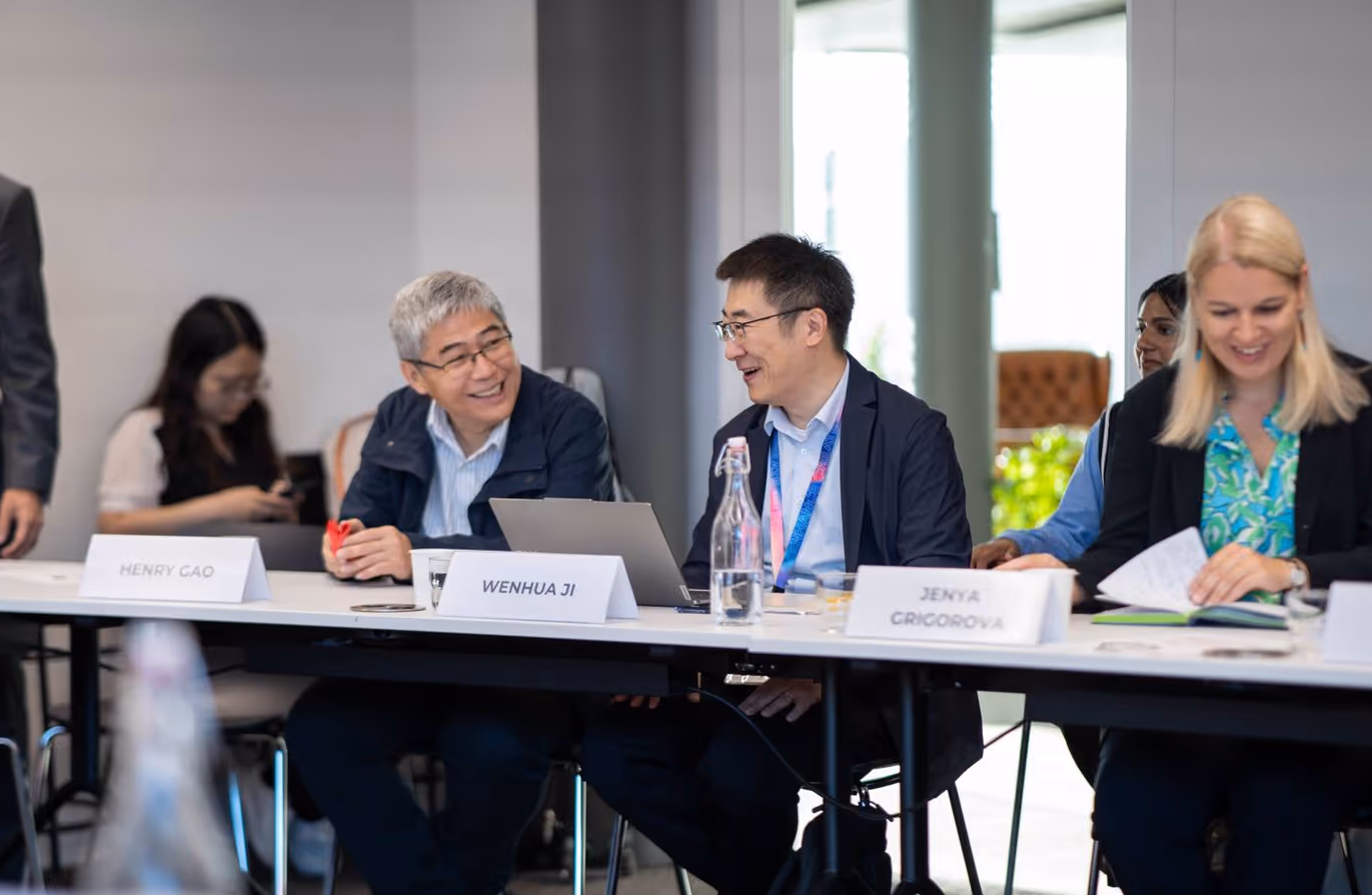 People sit at a conference table smiling, with name tags and laptops in a bright meeting room.