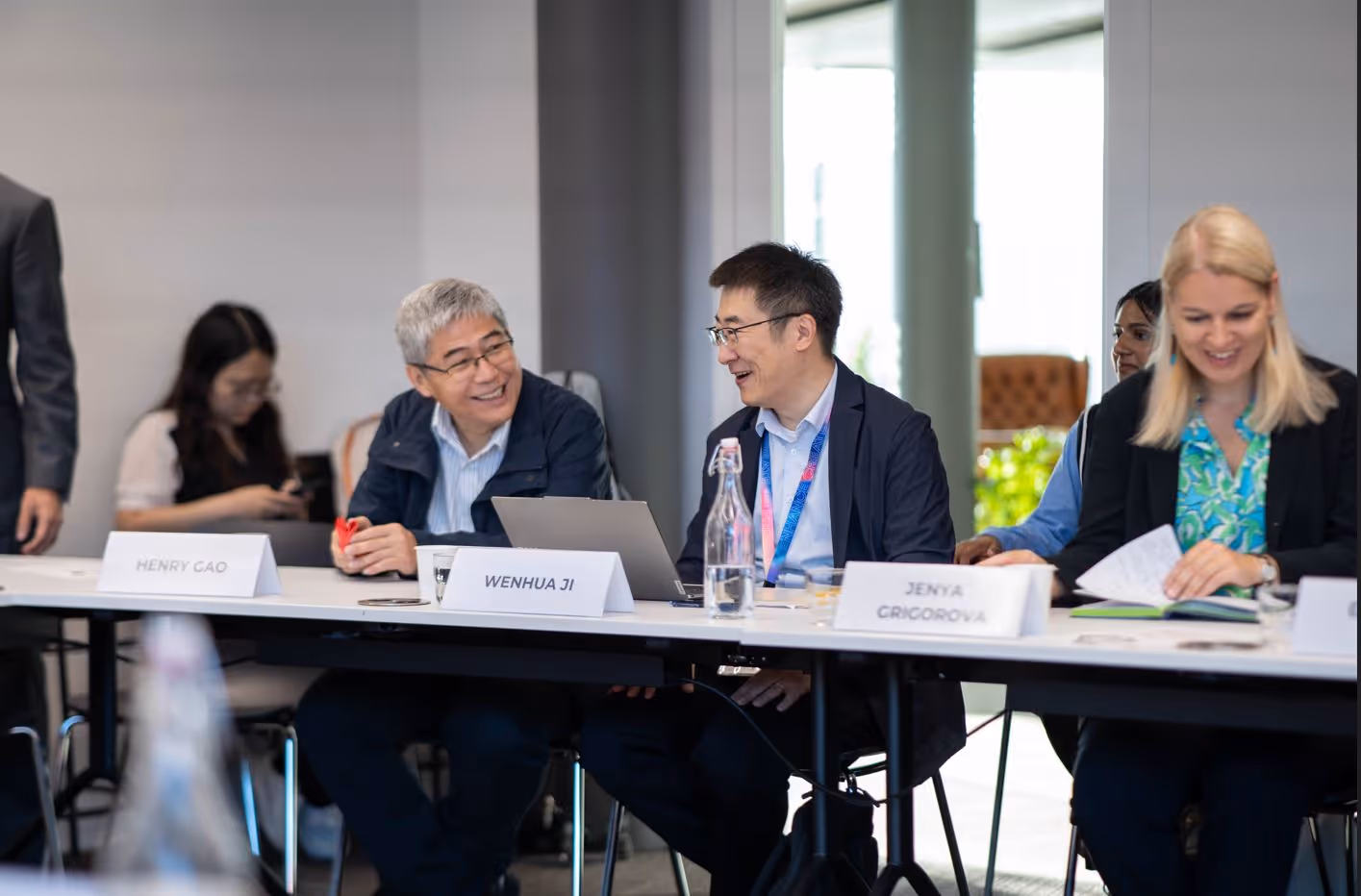 People sit at a conference table smiling, with name tags and laptops in a bright meeting room.