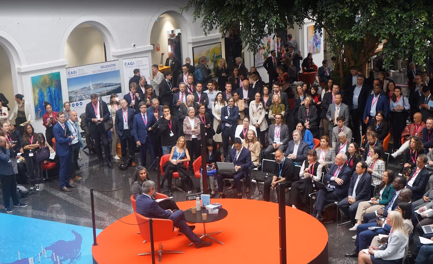 A large audience listens to a man speaking on a round red stage with two chairs and a table.