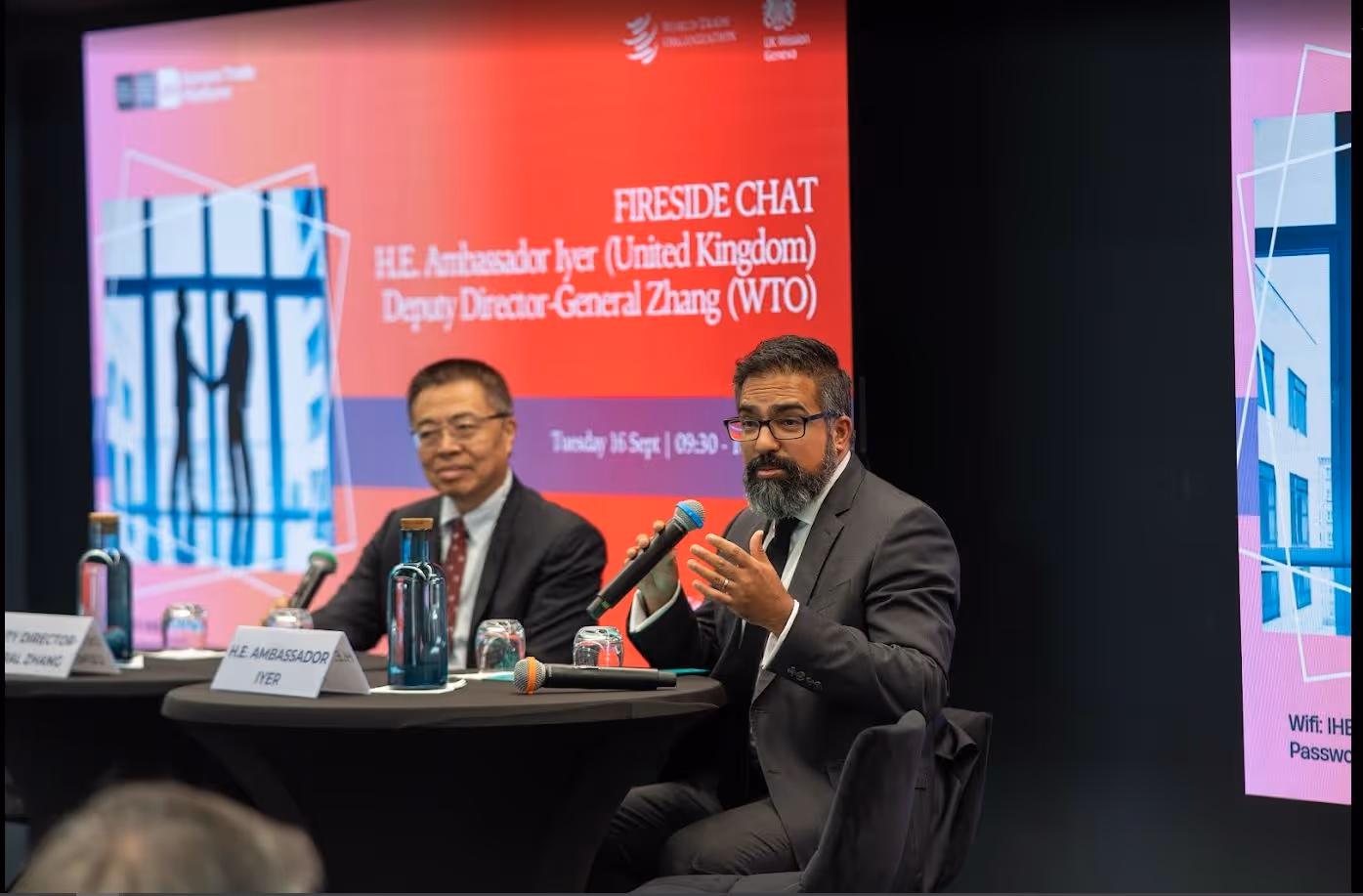 Two men in suits speaking at a fireside chat event with microphones and water bottles on the table.