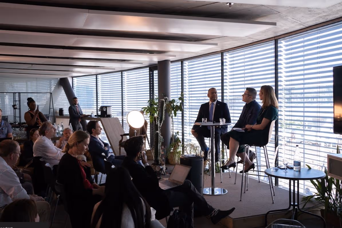 Three panelists seated on high chairs in front of an audience in a modern conference room.