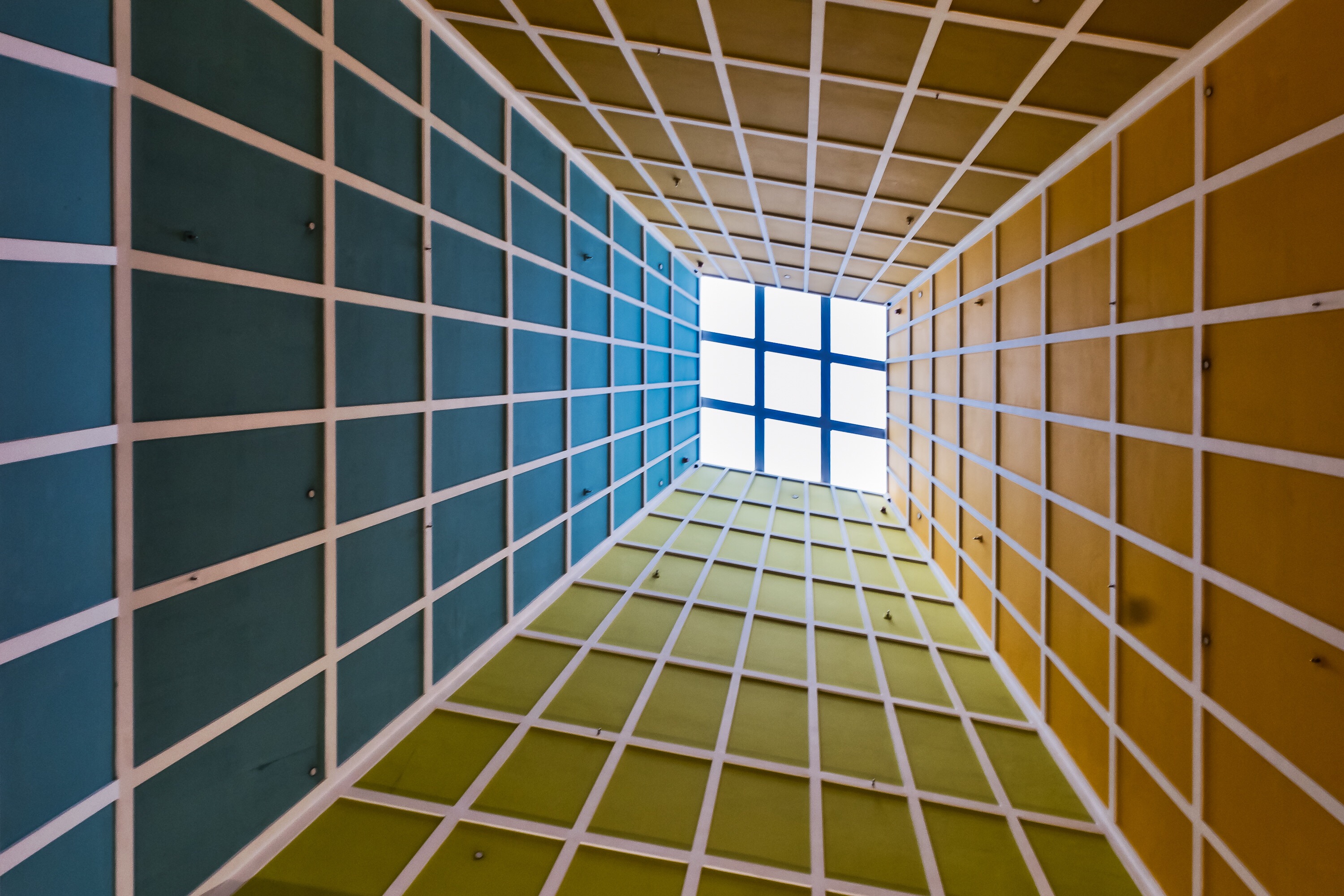 Skylight. Looking up through a geometric gridded structure toward a square of light — teal, yellow-green, amber panels converging on a bright opening. Conceptually perfect for a guide about moving from the indirect to the direct — the grid as the page, the light as the world beyond it. Off the Page
