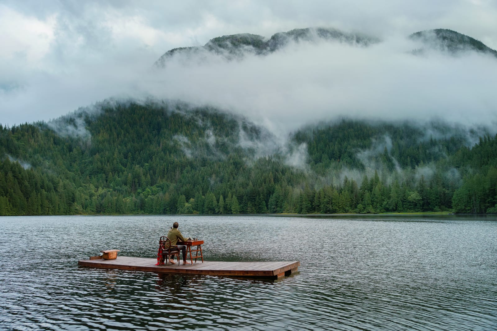 A man sitting barefoot at a table on a floating plaform in the middle of a lake with his back to the camera looking out at the mountains in front of him. Off-the-Grid
