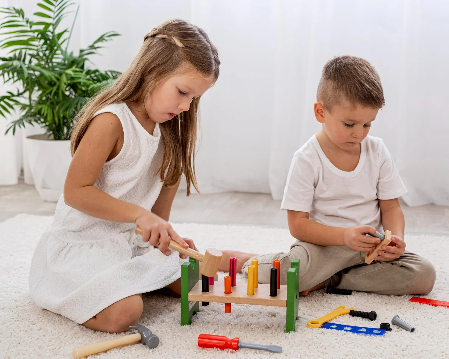 Girl and boy sitting on floor playing with toy tools and wooden peg board on a white rug.