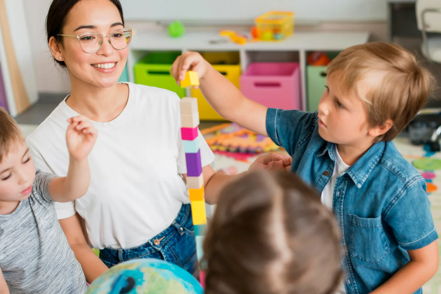 Teacher and children stacking colorful wooden blocks in a classroom with storage bins behind them.