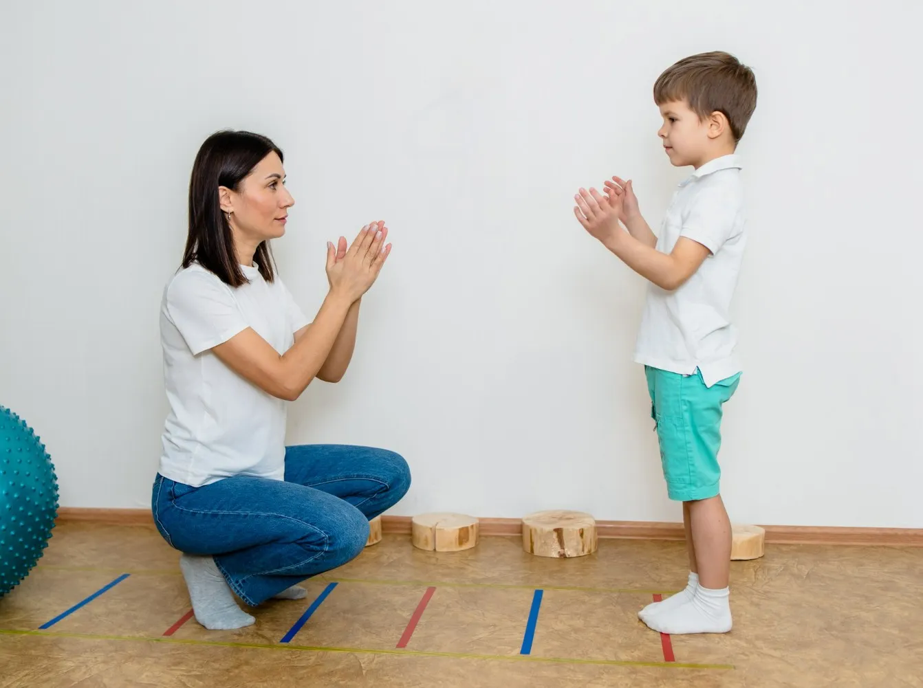 Woman squatting and clapping hands with young boy standing opposite her indoors.
