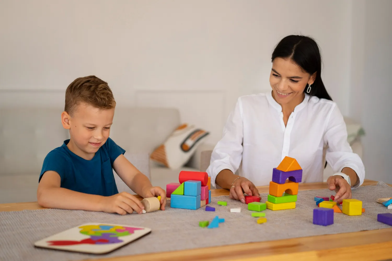 Woman and boy playing with colorful wooden blocks and a puzzle on a table indoors.