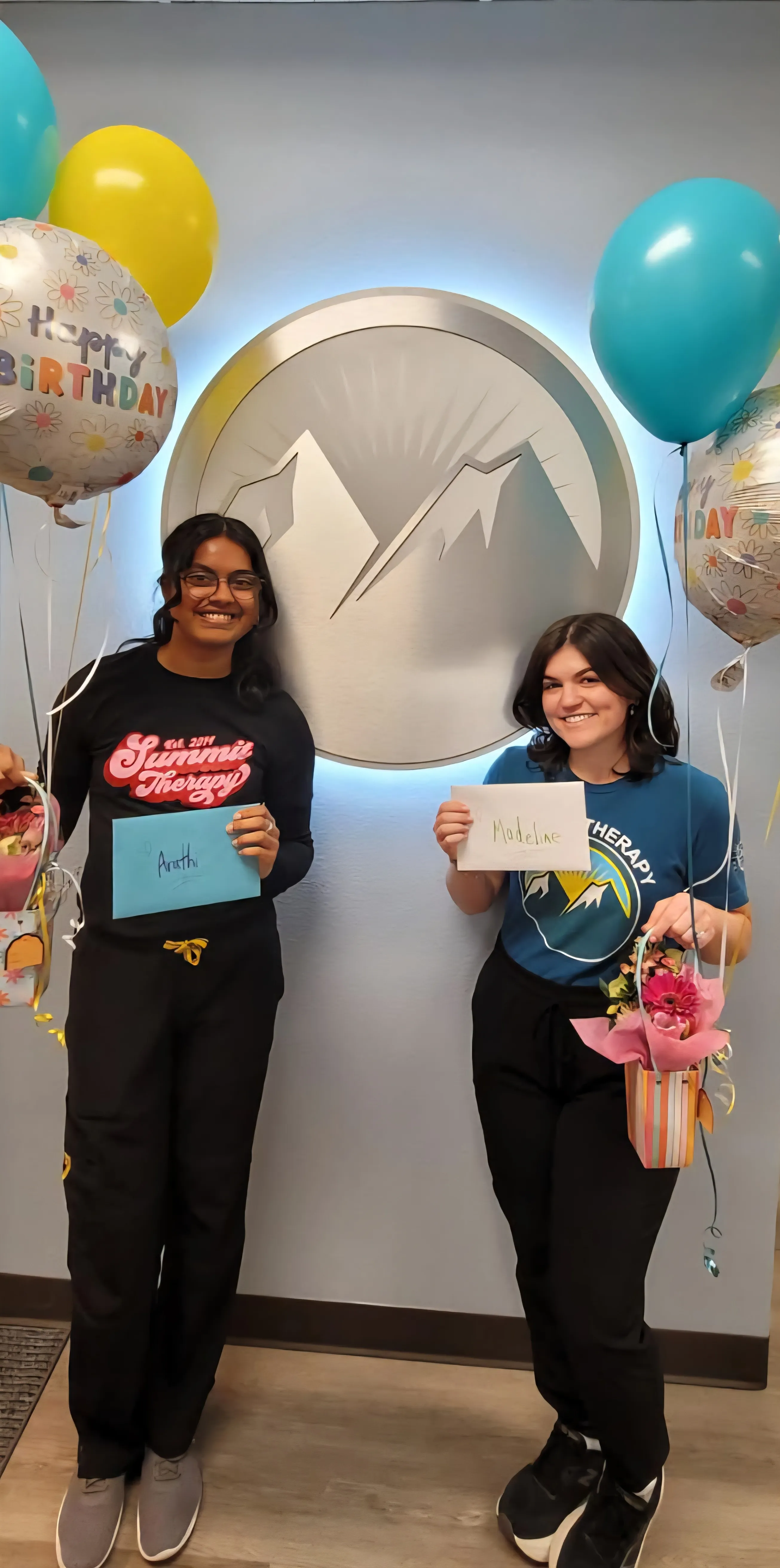 Two smiling women holding balloons, flowers, and cards with names Anathi and Madeline in front of a mountain logo.