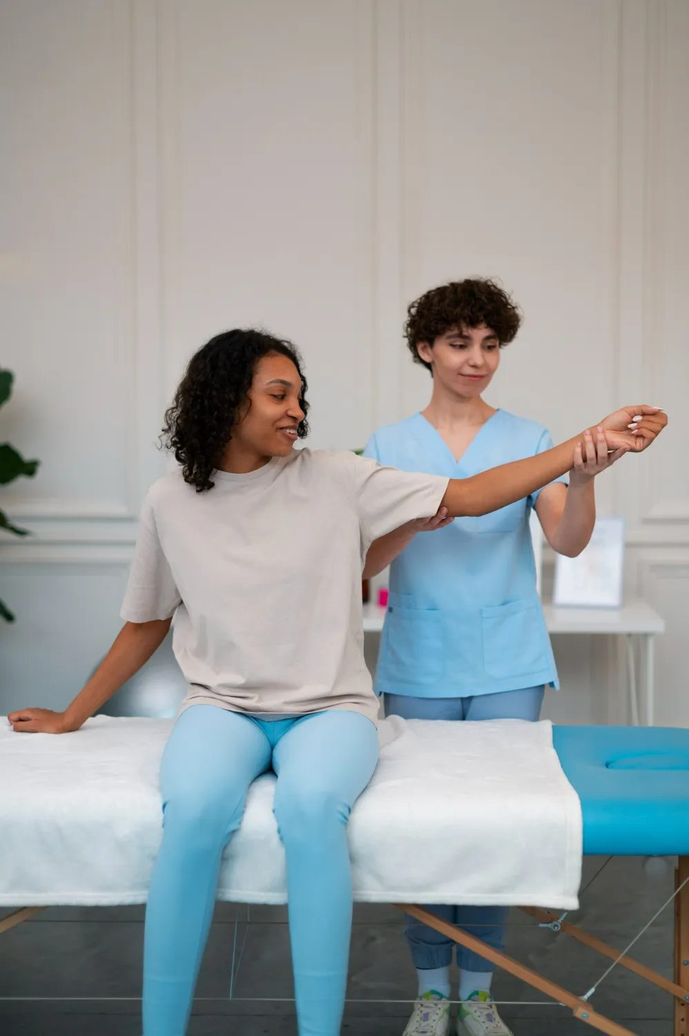 Therapist in blue scrubs assisting a woman stretching her arm sitting on a white massage table.