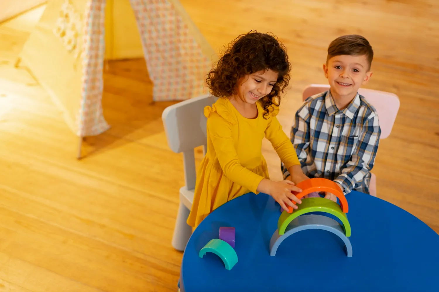 Two children smiling and playing with colorful wooden arches at a blue table in a playroom.