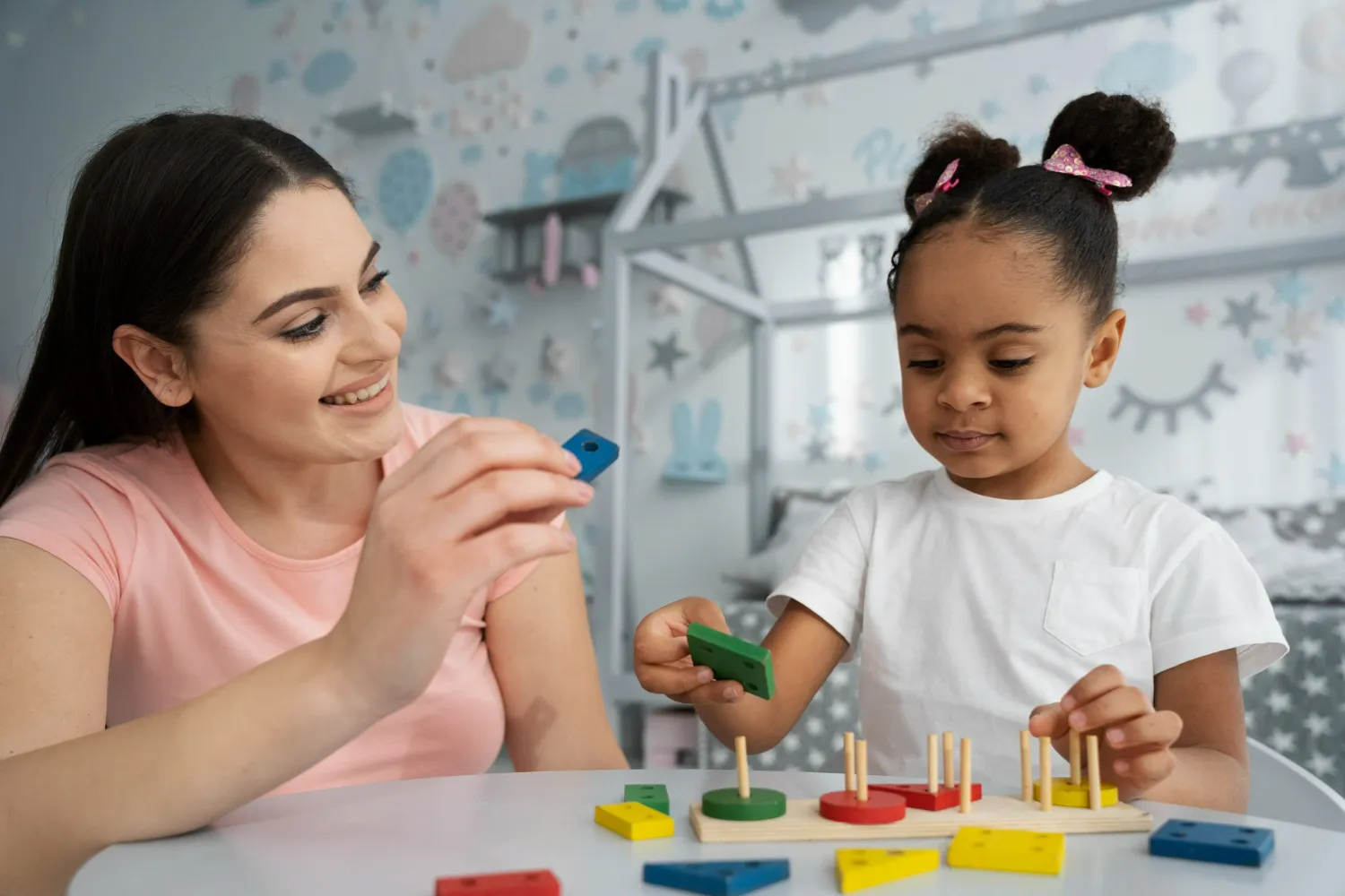 Smiling woman playing with a child using colorful shape sorter toy at a white table.
