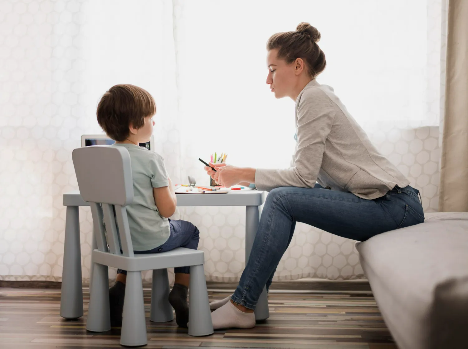 Woman helping a young boy with drawing at a small table in a bright room with wooden floor.