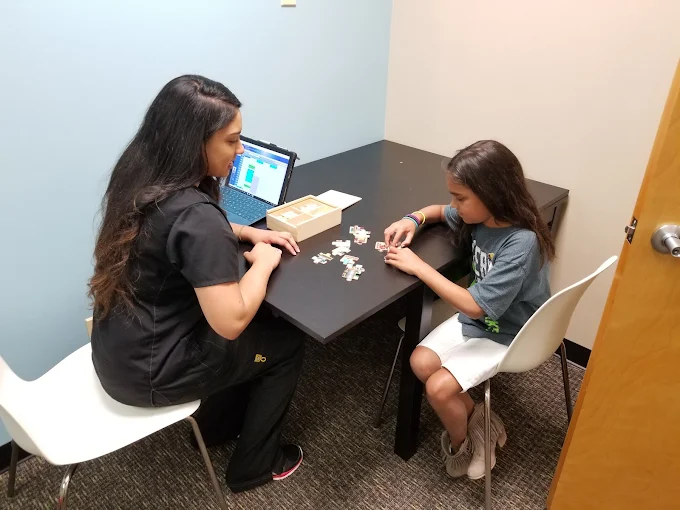 Woman and child seated at a table working on a puzzle with a laptop open nearby.