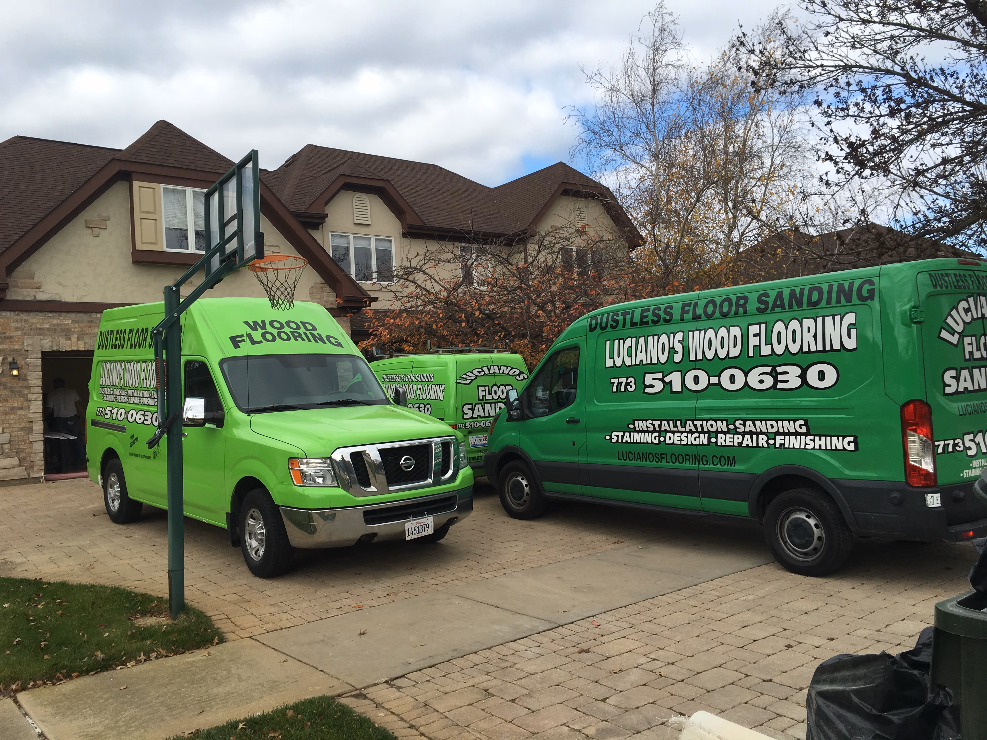 Two bright green vans for Luciano's Wood Flooring parked in a residential driveway with basketball hoop nearby.