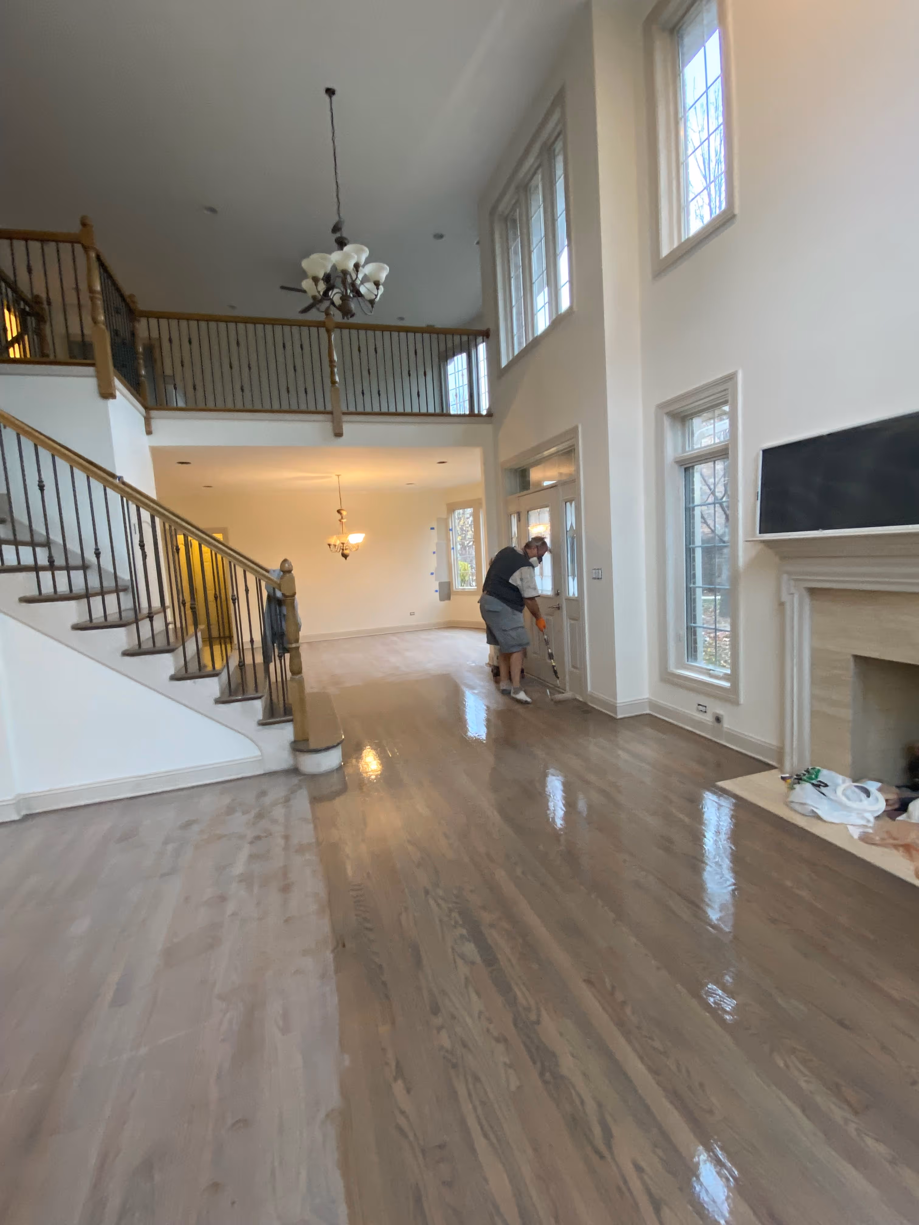Person applying finish on hardwood floor in large two-story room with staircase and fireplace.