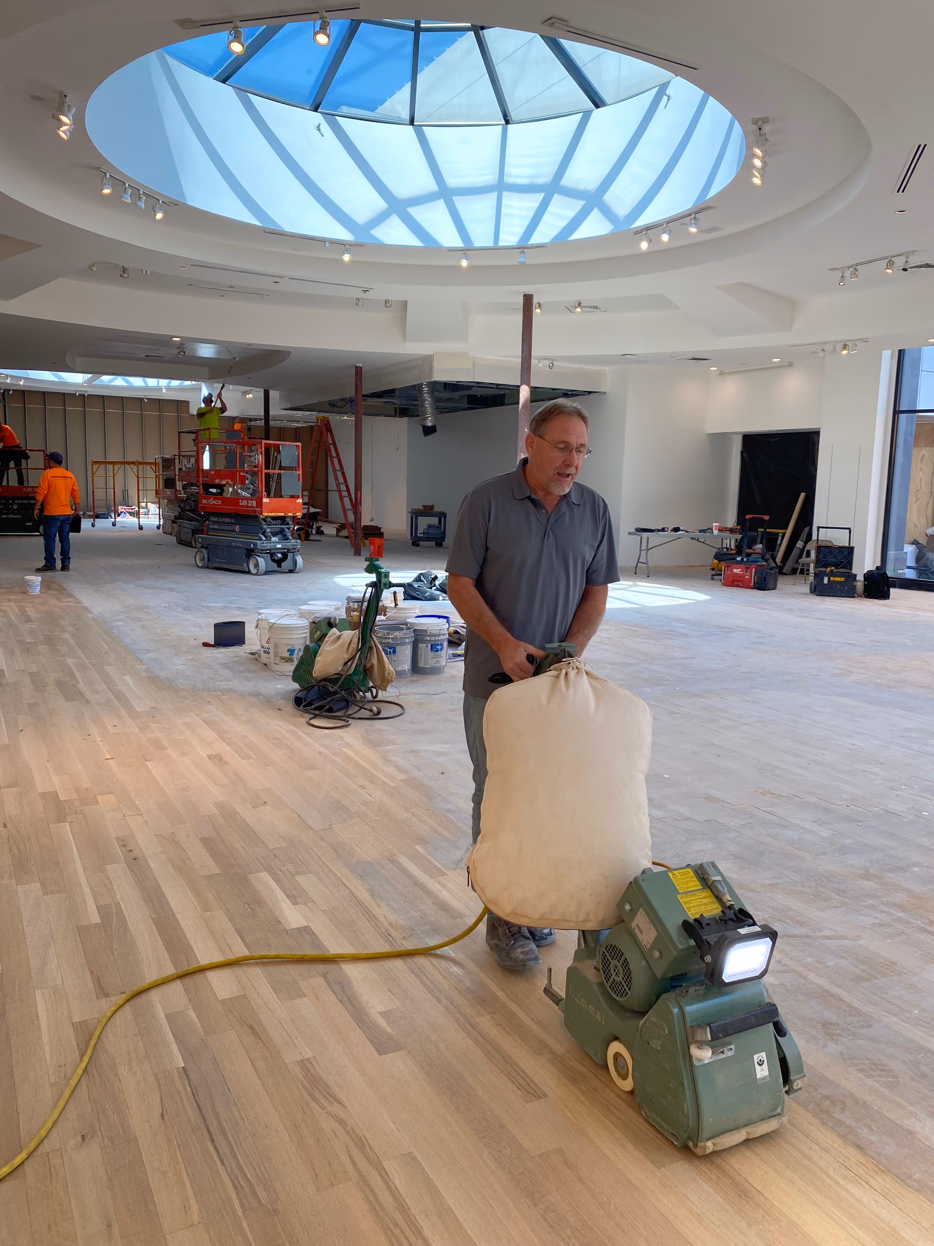 Man operating a floor sanding machine on wooden floor under a large skylight in a building under construction.