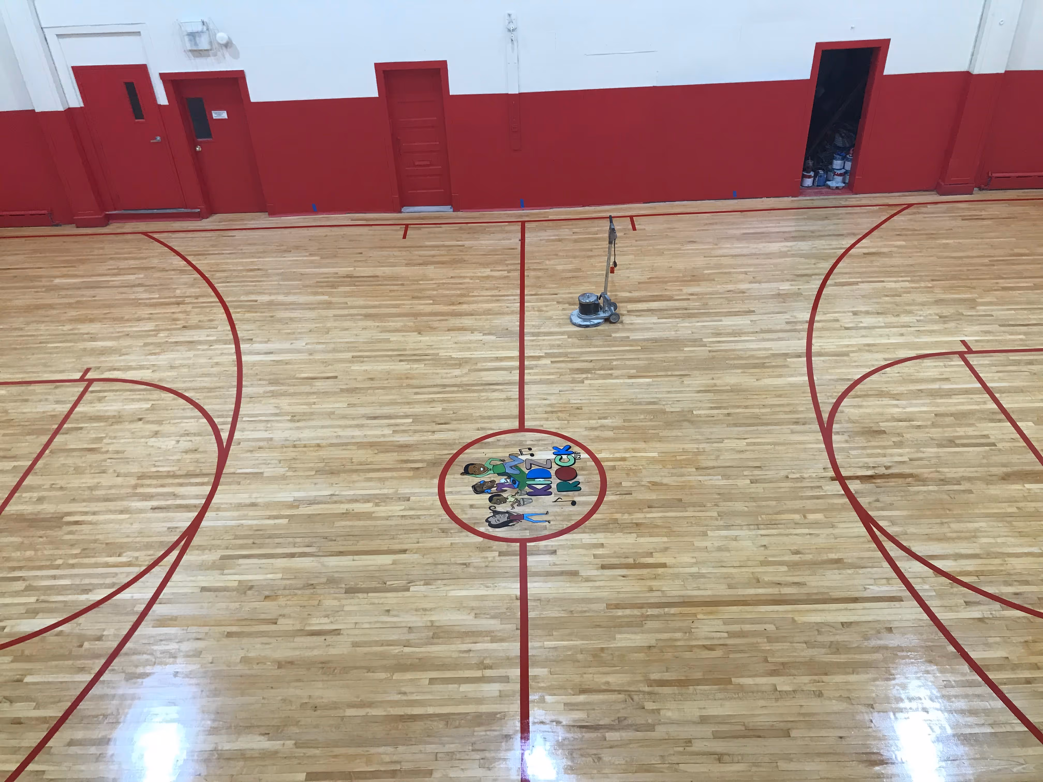 Indoor basketball court with red lines and a colorful Kids Rock logo at center court.
