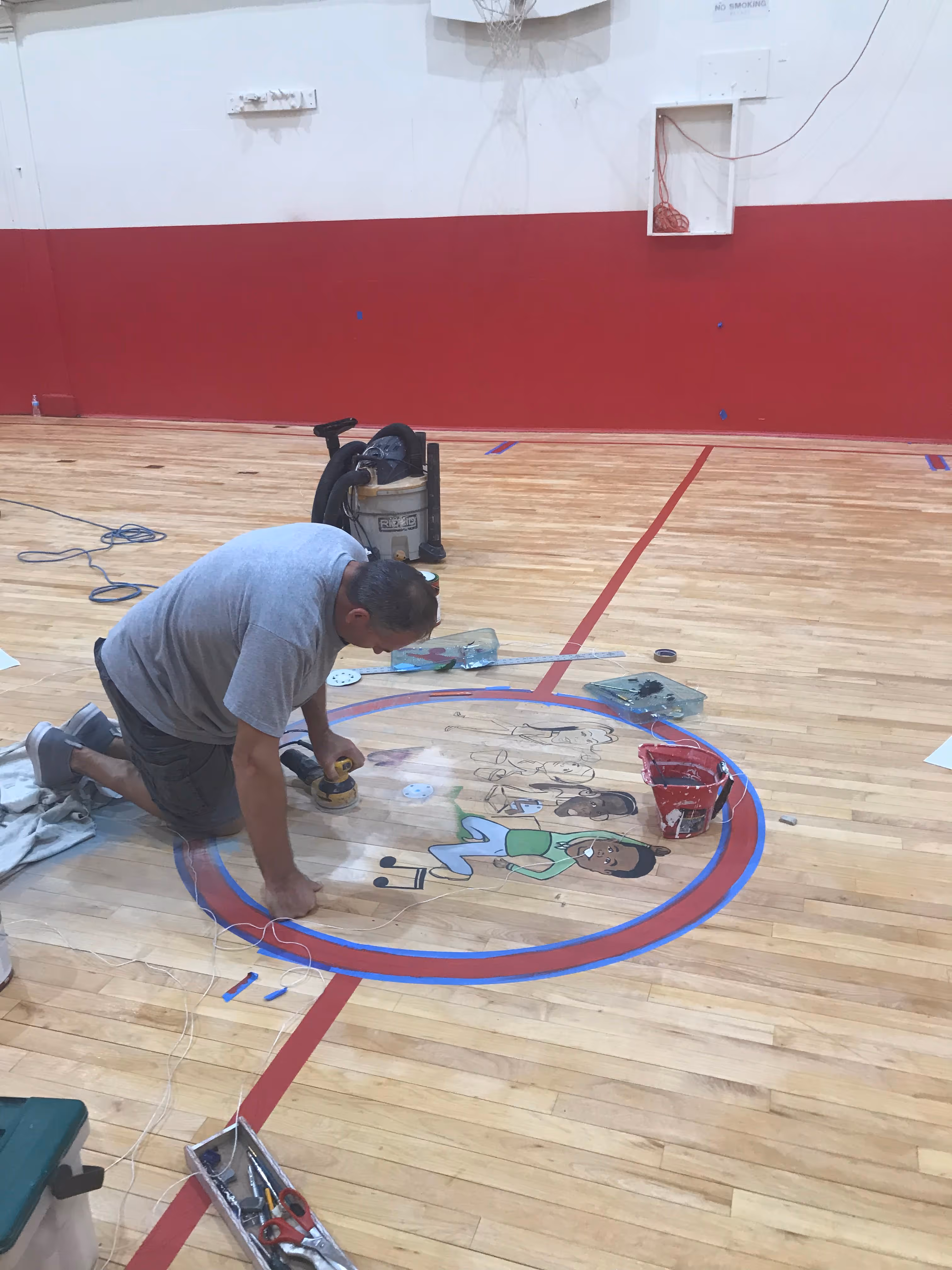 Man sanding a cartoon mural painted on the center court of a wooden gym basketball floor.