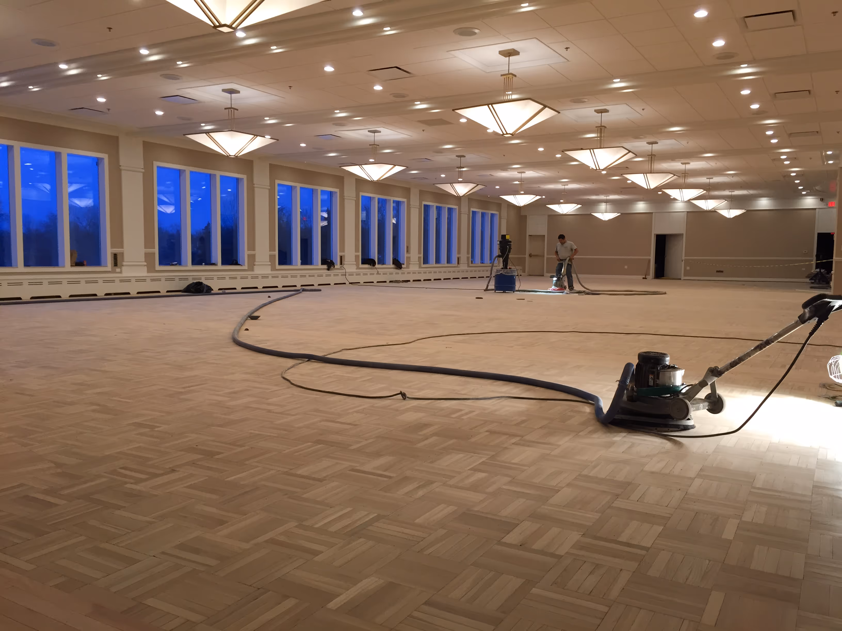 Large empty room with parquet floor being sanded by a worker using floor sanding machines.