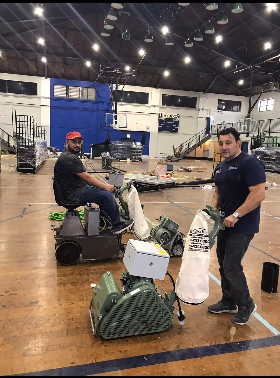 Two men with floor sanding machines on the wooden floor of a gymnasium with blue and white walls.