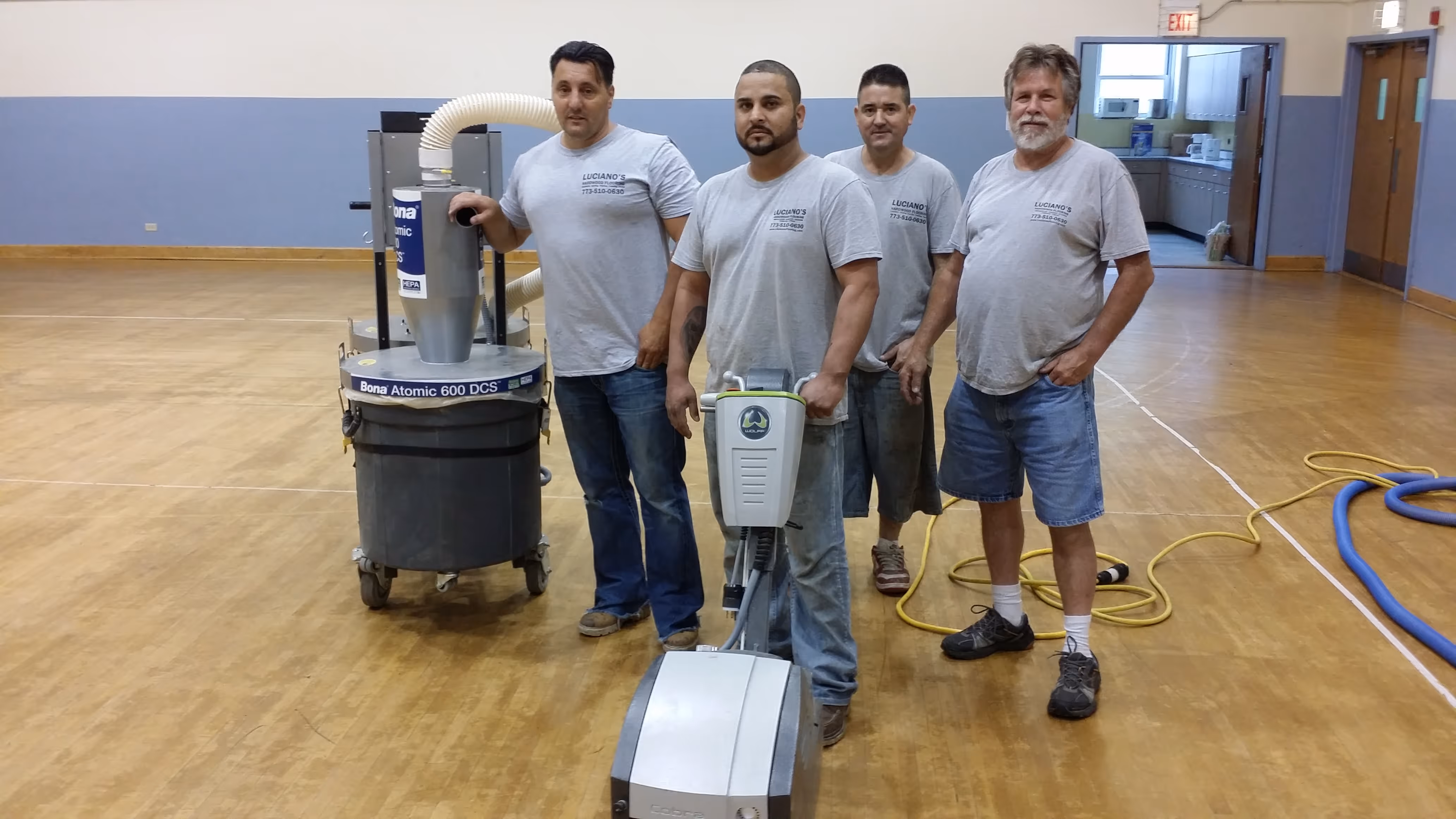 Four men in gray shirts standing on a wooden floor with floor sanding machines in a gym.