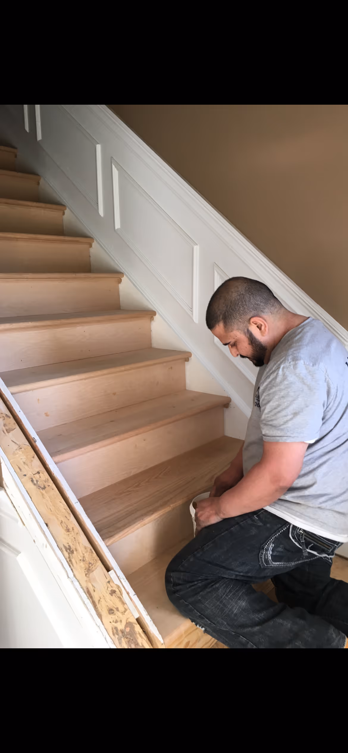Man kneeling on wooden stairs installing or working on the steps near a white paneled wall.