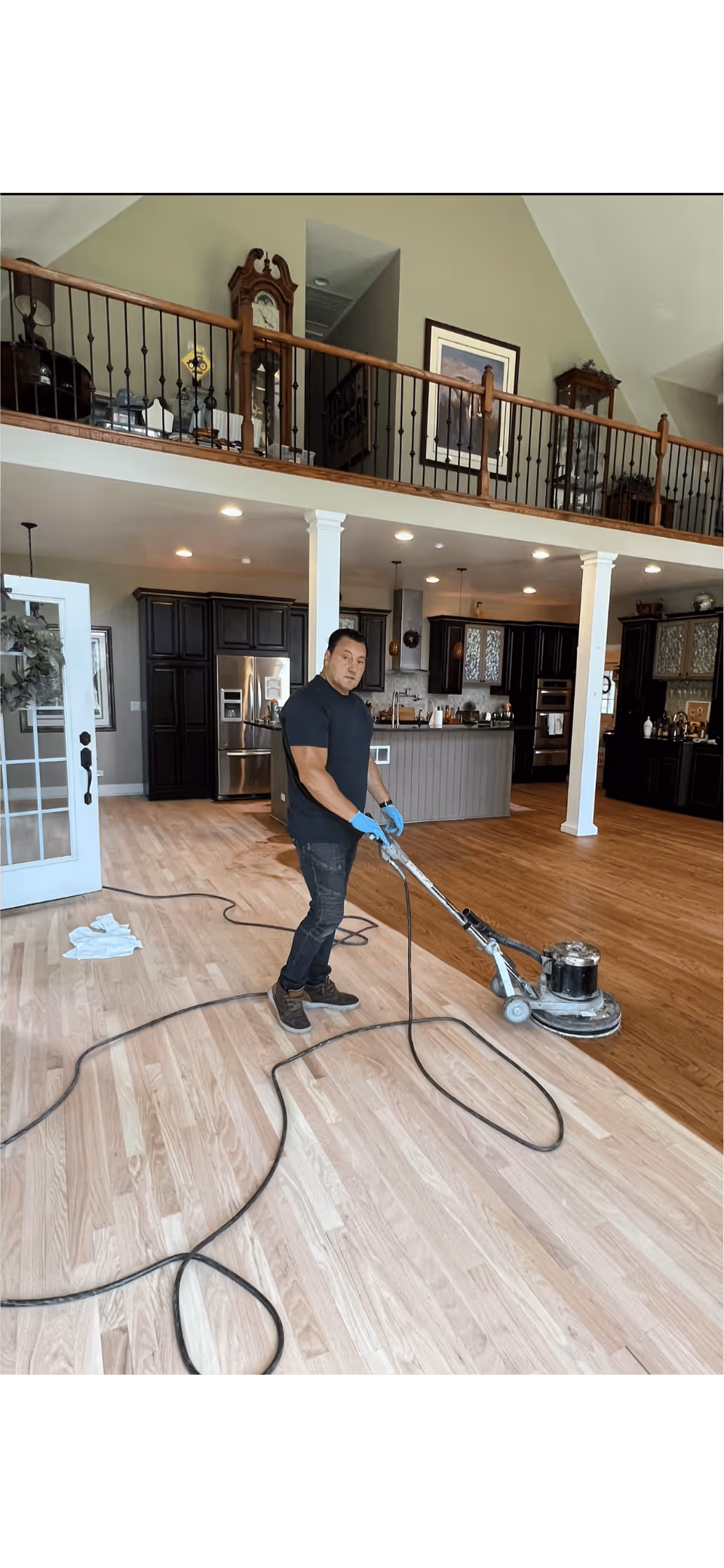 Man with blue gloves using floor polishing machine on hardwood in a large modern kitchen.
