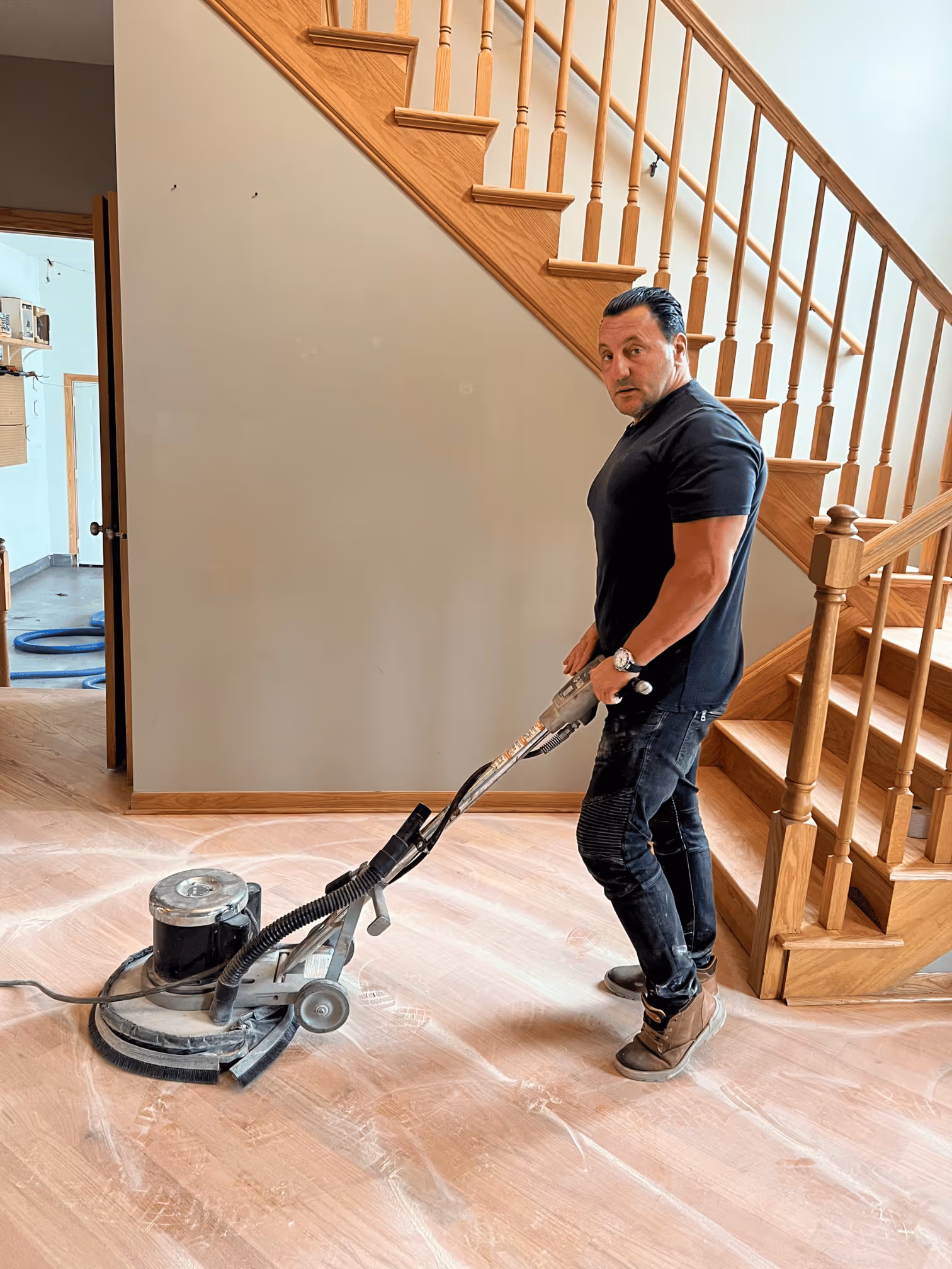 Man using a floor buffer to polish hardwood floors near a wooden staircase inside a house.